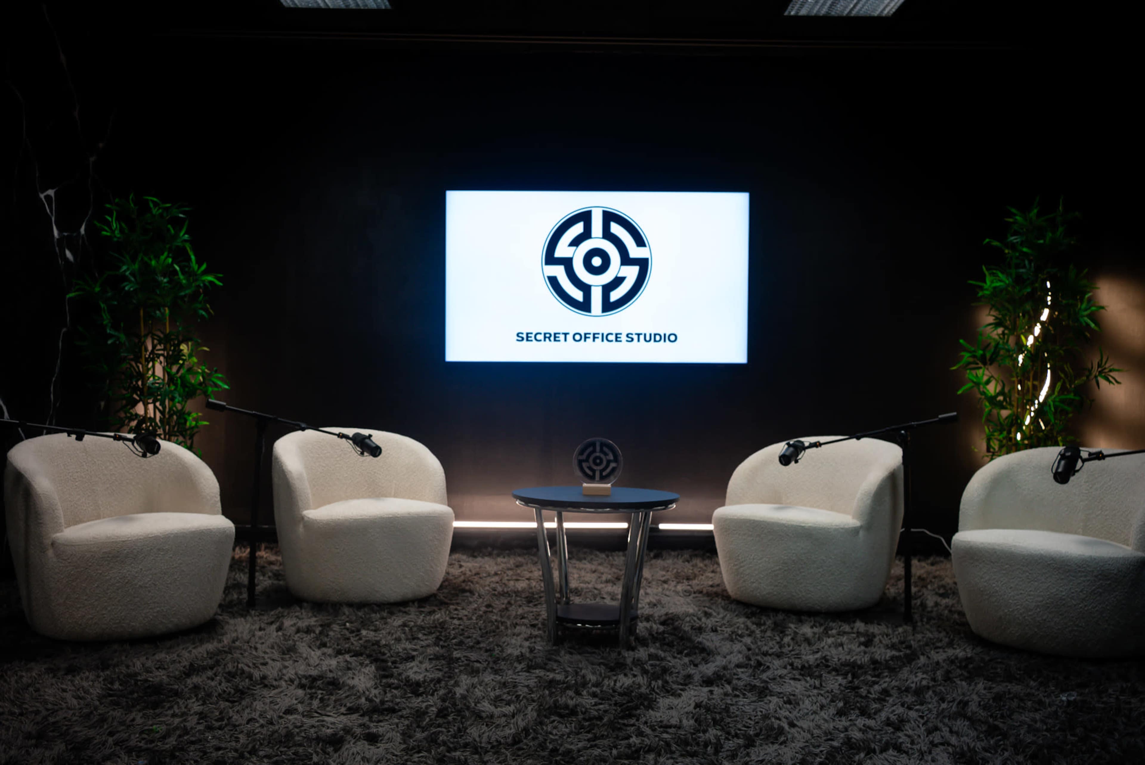 The image features a modern studio setup with four white chairs arranged around a central table, located beneath a screen displaying the logo "SECRET OFFICE STUDIO" against a dark background.