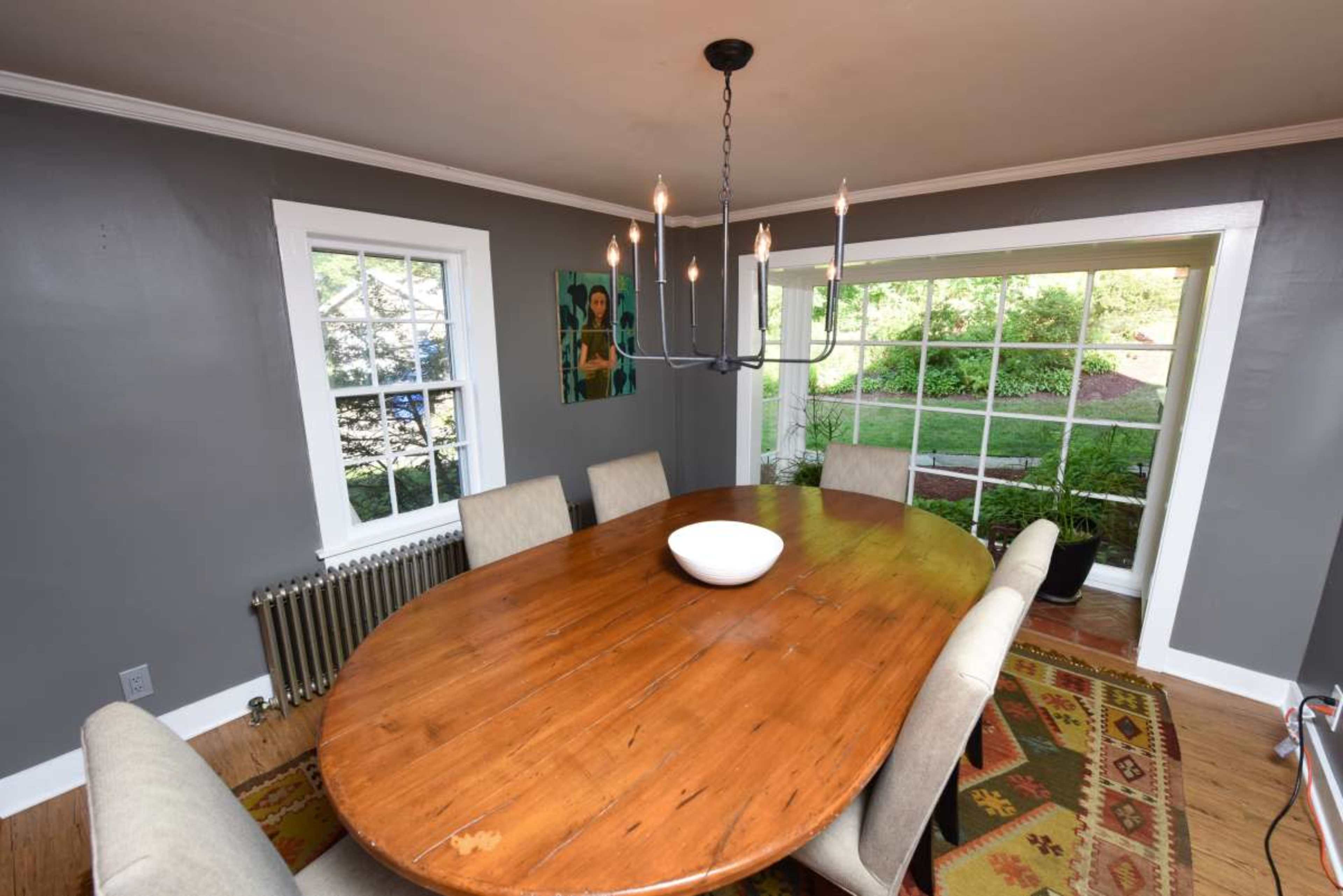 A dining room with a large oval wooden table surrounded by beige chairs, a modern chandelier overhead, and large windows that provide a view of greenery outside.