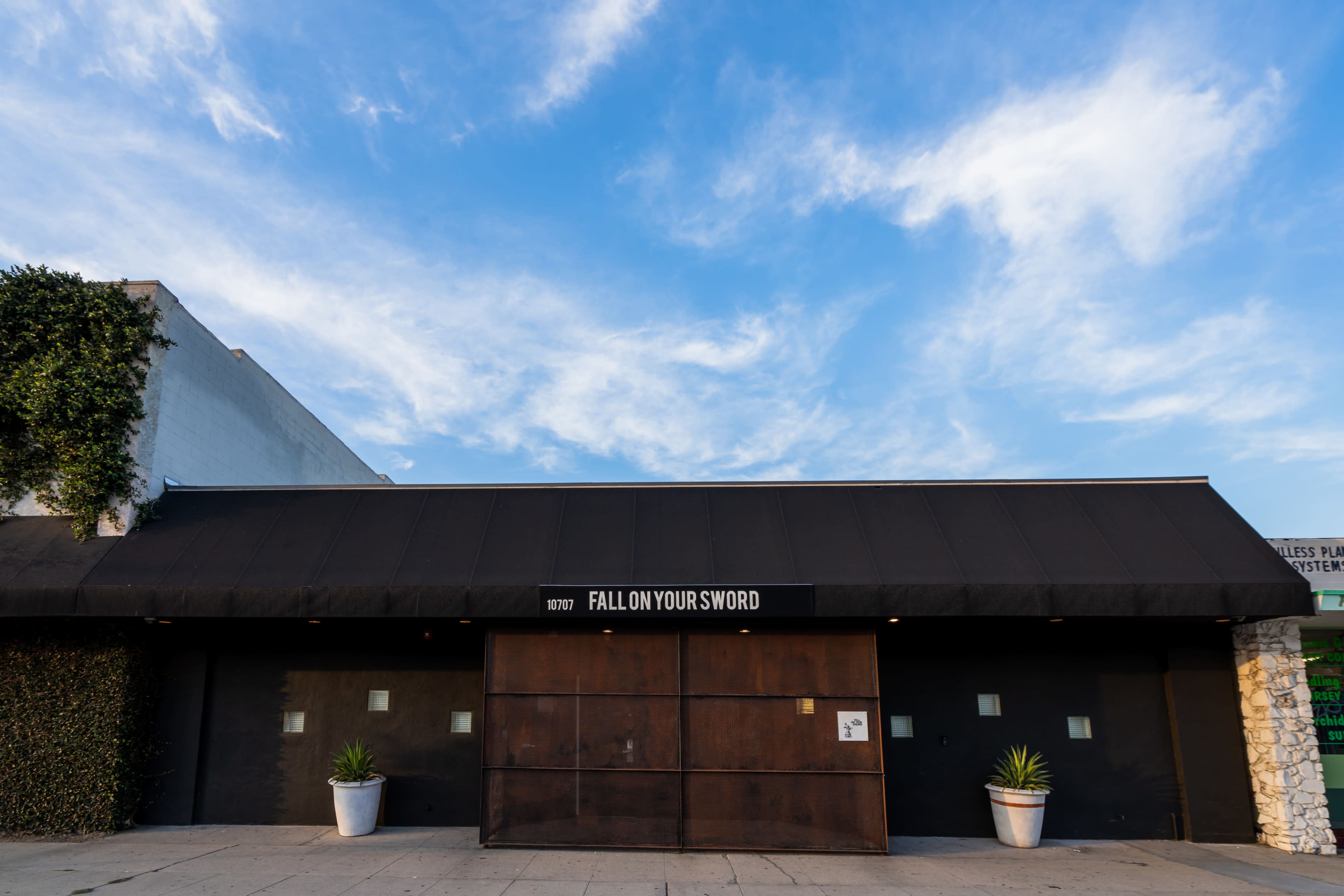 The exterior of a building with the words "FALL ON YOUR SWORD" displayed above a large wooden entrance, set against a clear blue sky.