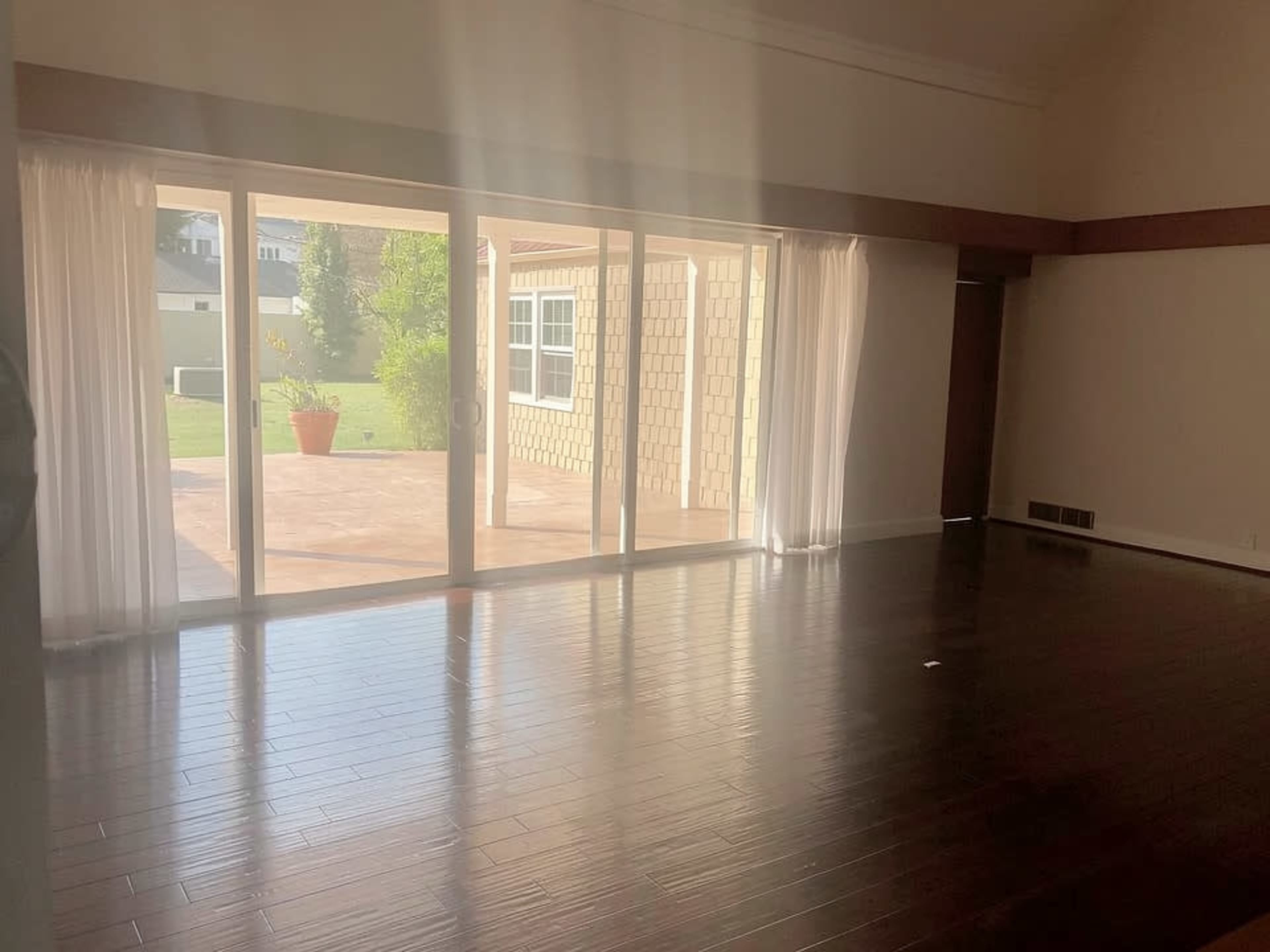 The image shows a spacious, sunlit room with large glass doors opening to a patio area, featuring wooden floors and sheer curtains.