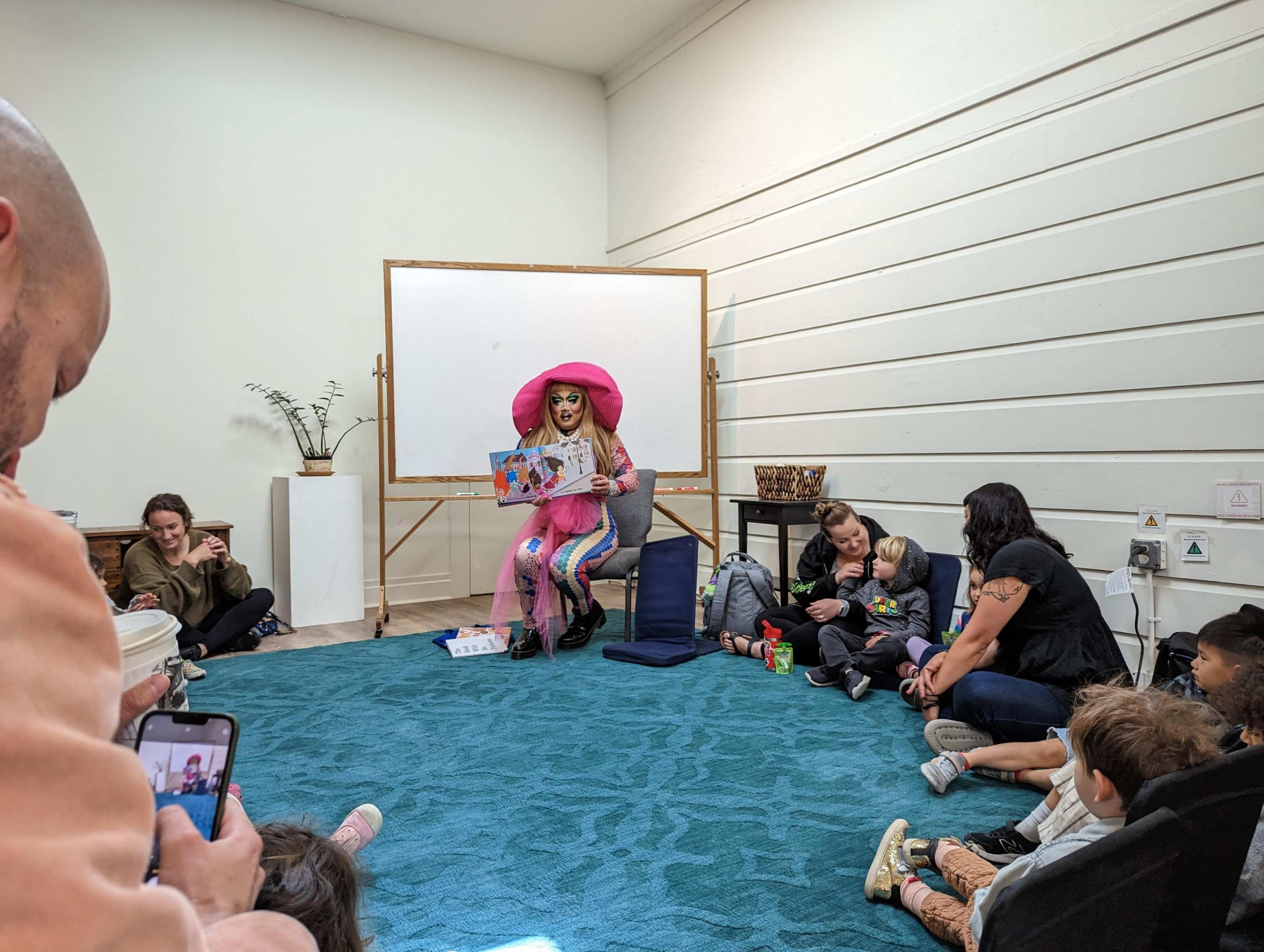 A person in colorful attire reads a story to a group of children and adults seated on the floor in a small, light-colored room.