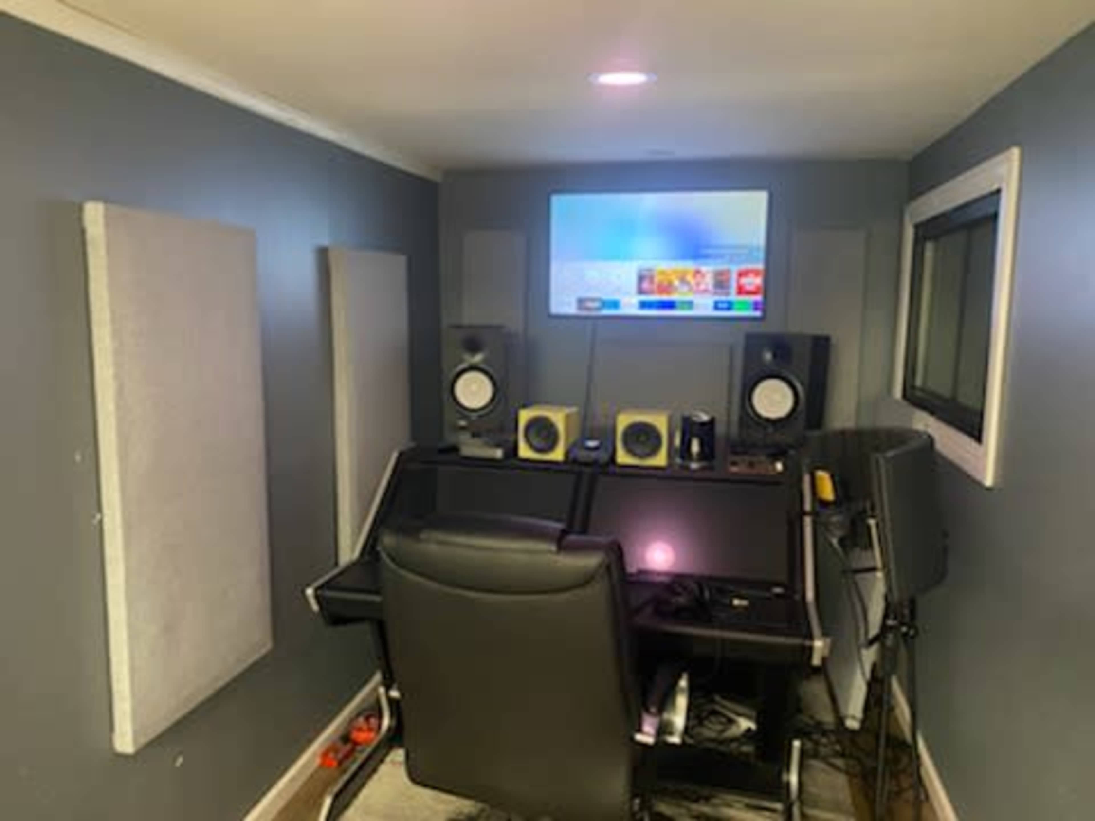 A small music studio with a desk, dual monitors, speakers, and soundproofing panels on the walls.