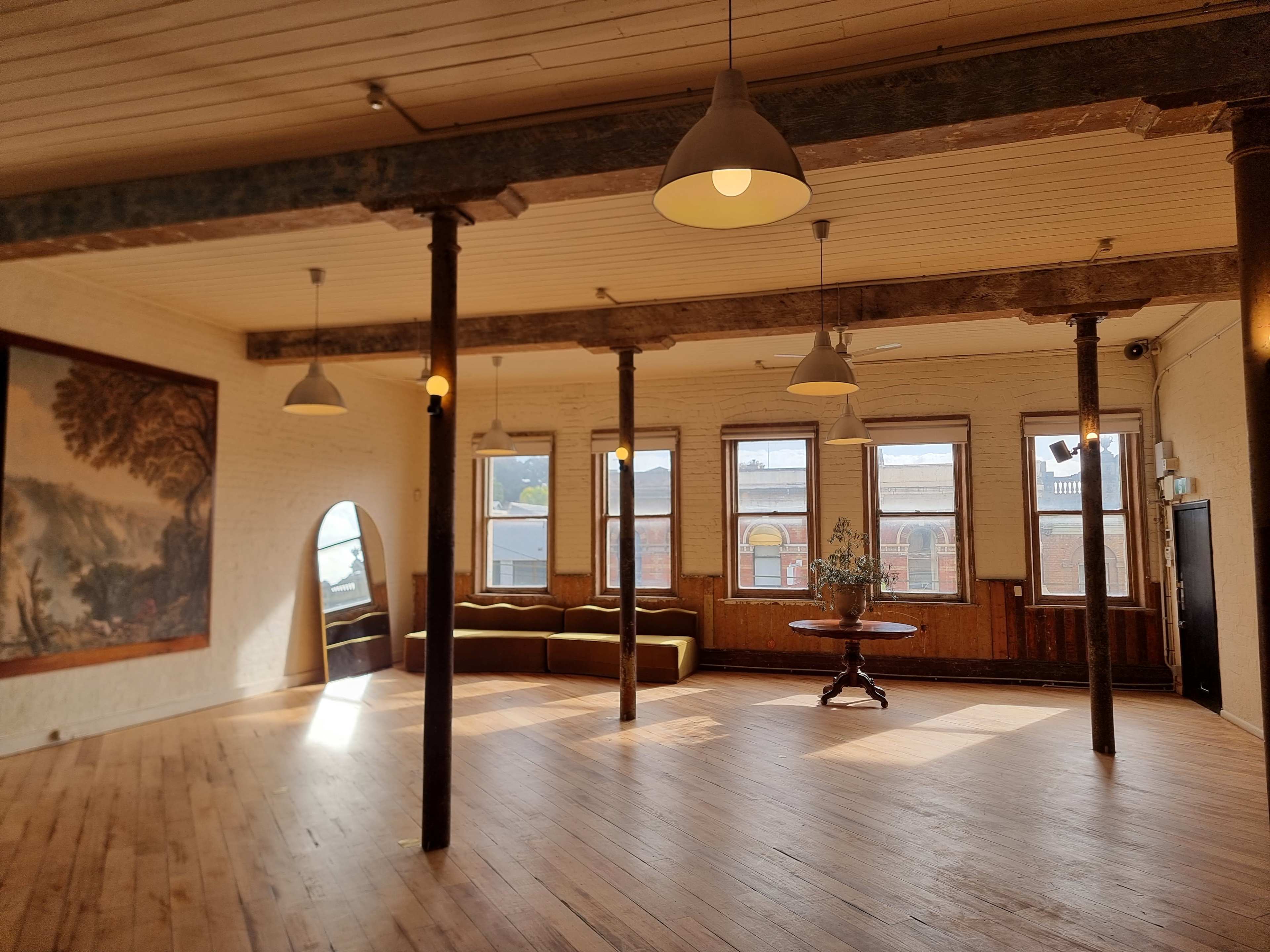 The image shows a spacious room with wooden floors, high ceilings, and large windows, featuring a decorative wall painting and a circular table surrounded by a couch.
