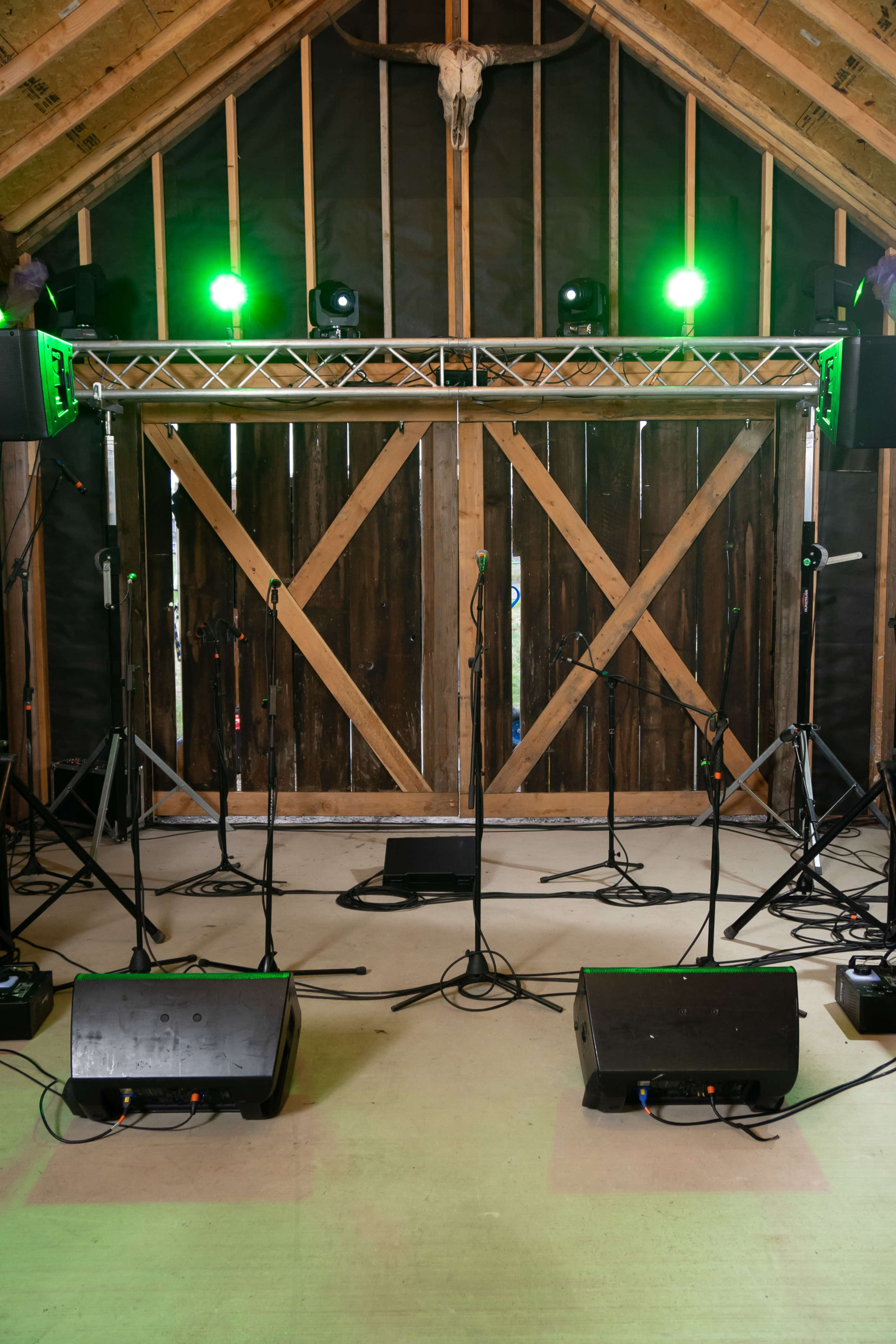 A stage setup with microphones and speakers is positioned in front of a large wooden door in a barn, illuminated by green lights.