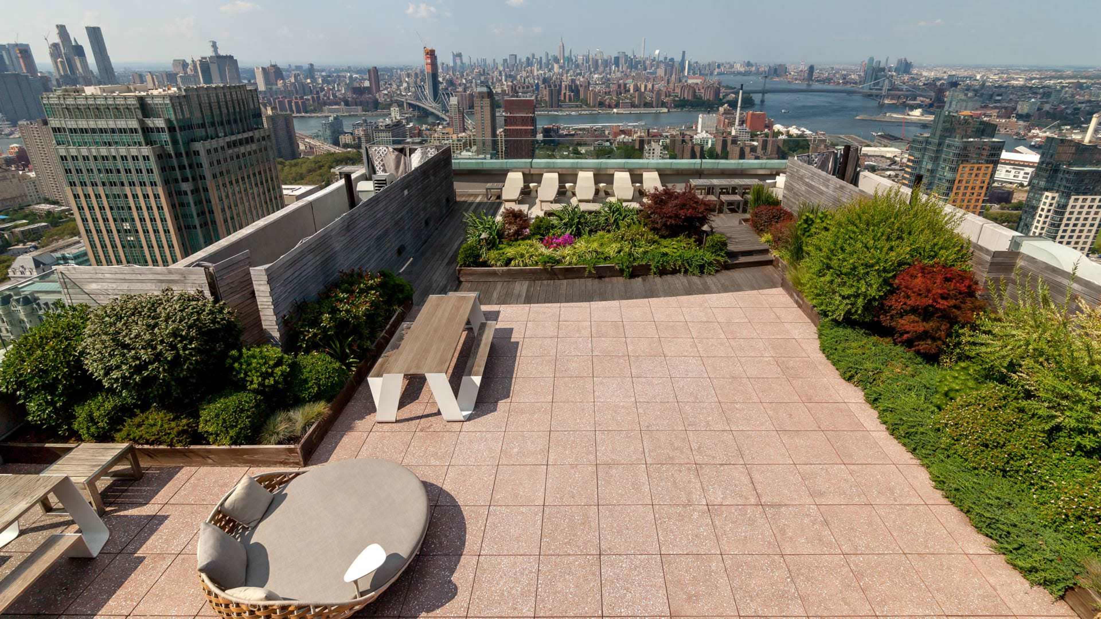 A rooftop terrace with a view of a city skyline, featuring landscaped greenery and outdoor furniture arranged on a tiled surface.