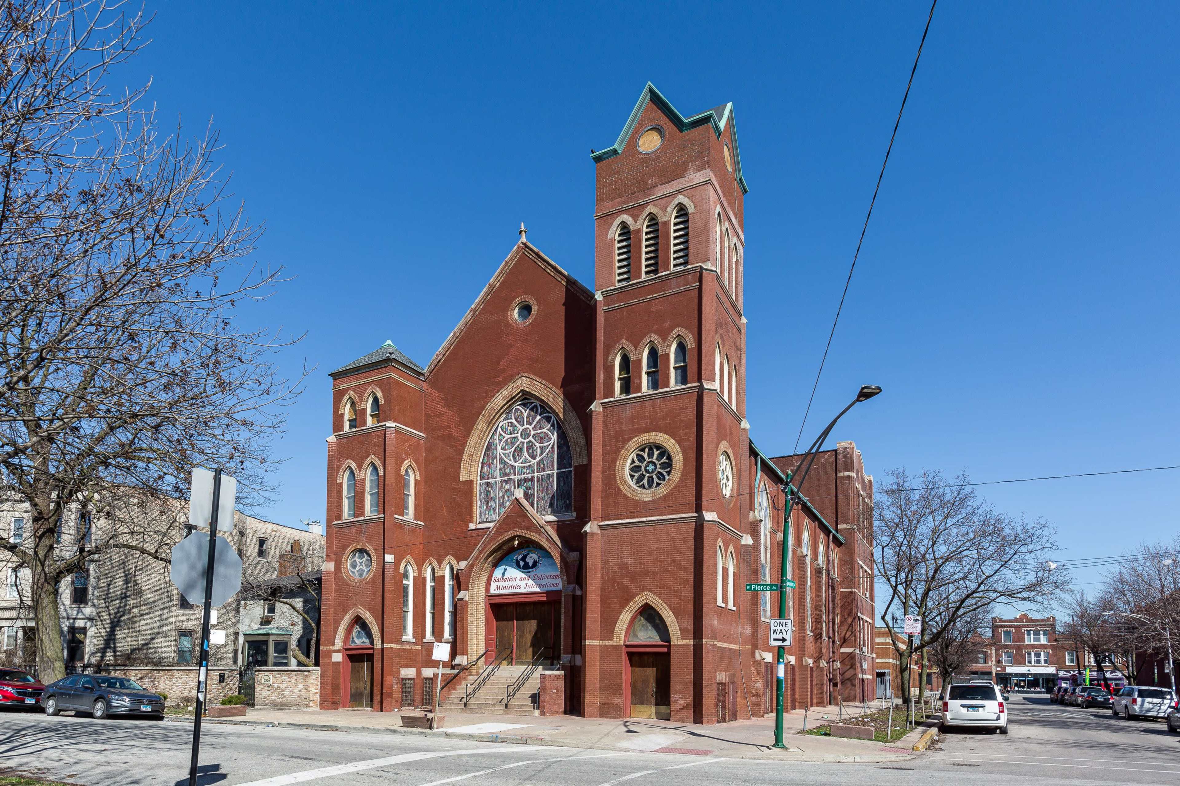 A large red brick church with a prominent rose window and a tall bell tower, set against a clear blue sky.