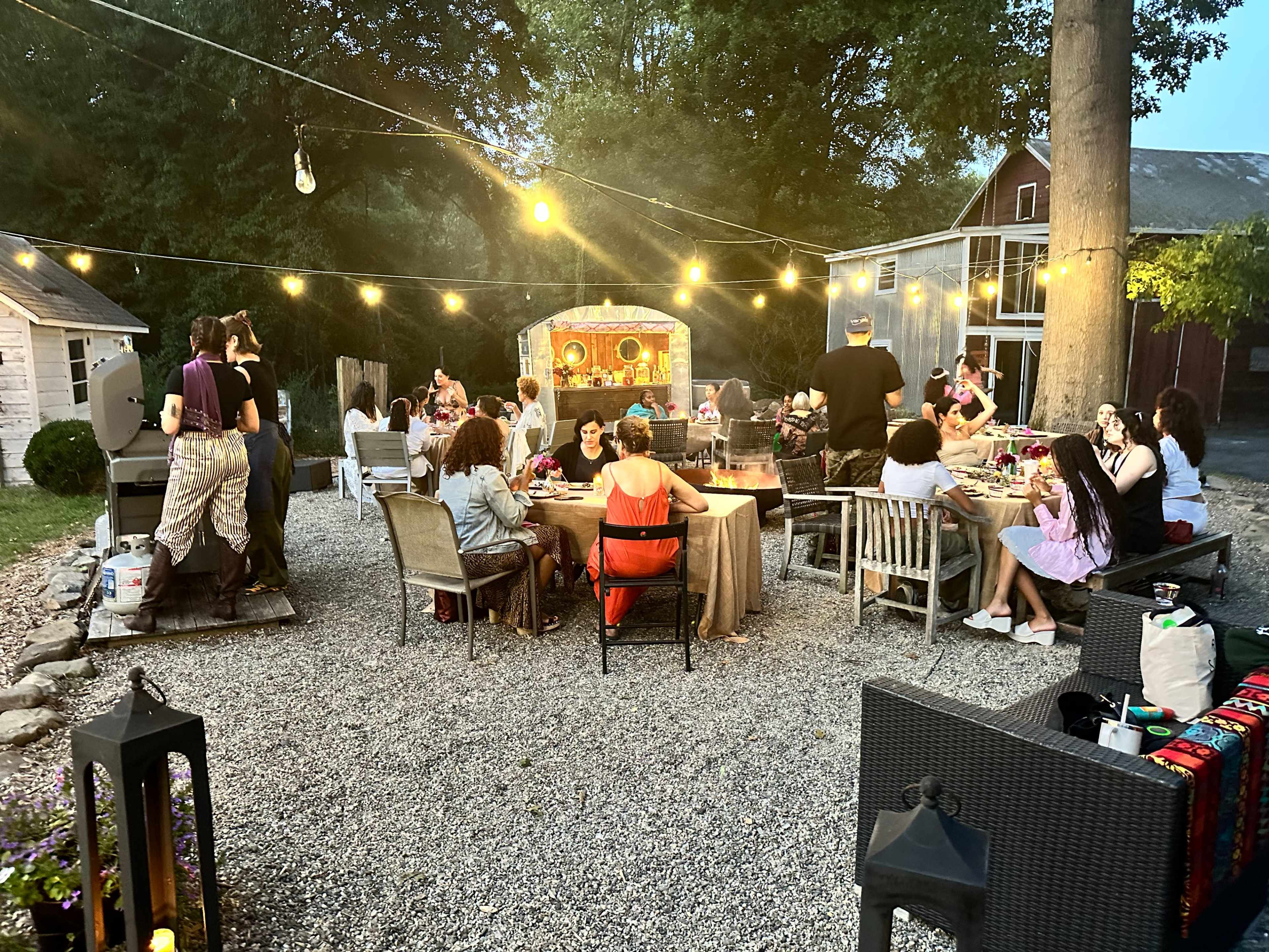 A large group of people gathers around a well-decorated outdoor table with string lights overhead, enjoying a meal in a gravel courtyard.