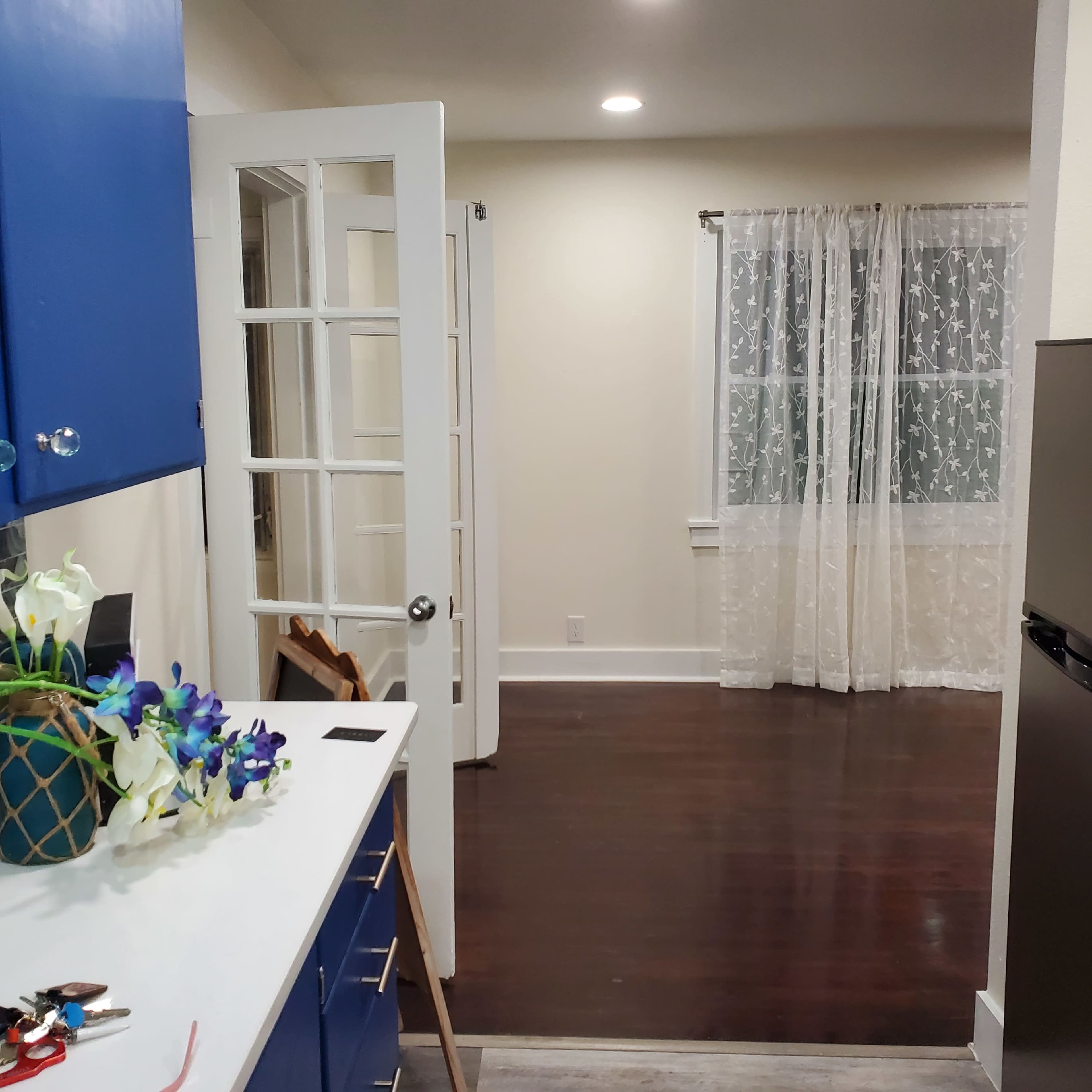 A view from a kitchen with blue cabinets leading into a room with a window covered by a sheer curtain.