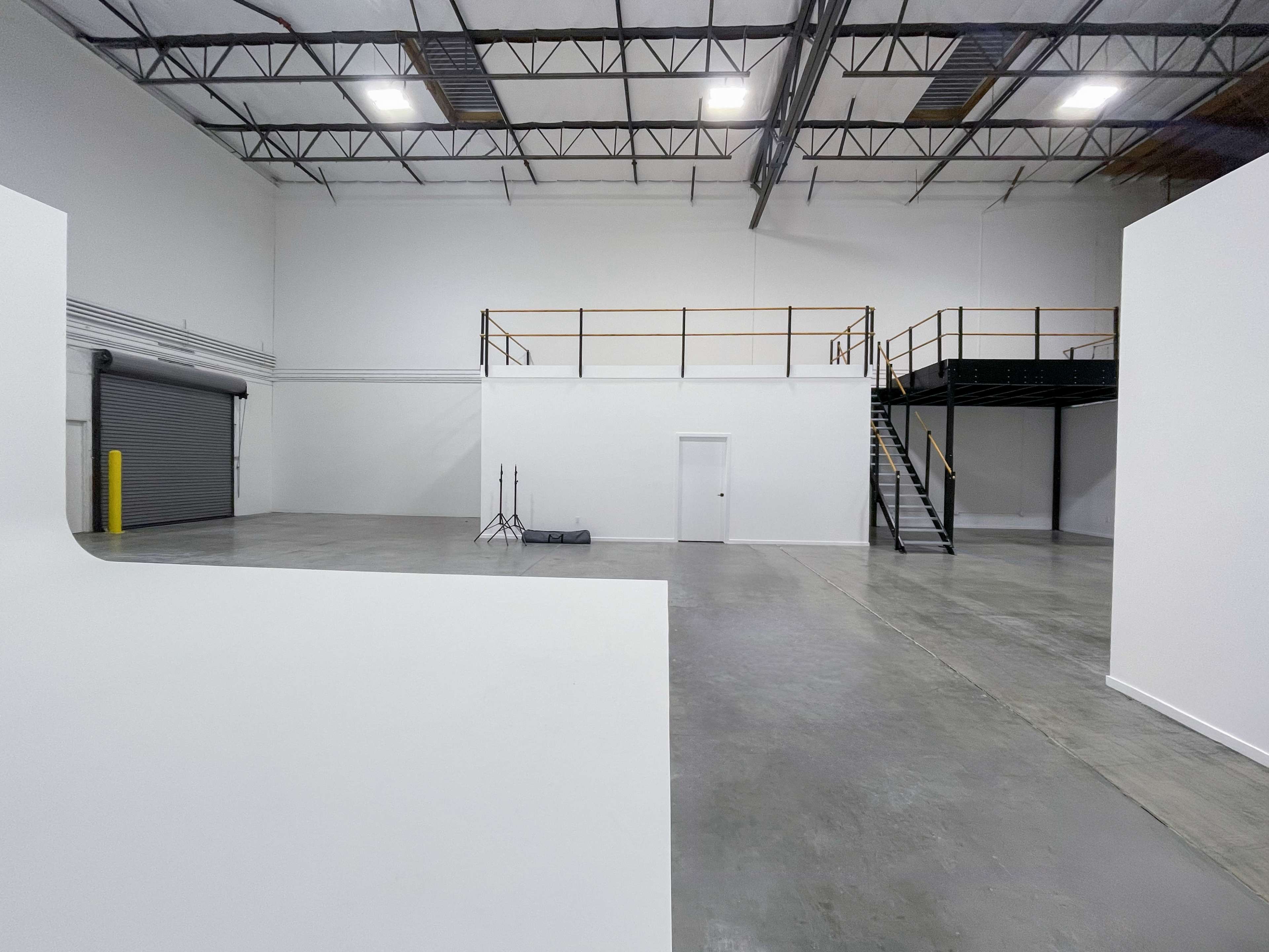 The image shows a large, empty warehouse space with white walls, a concrete floor, and a mezzanine railing in one corner.