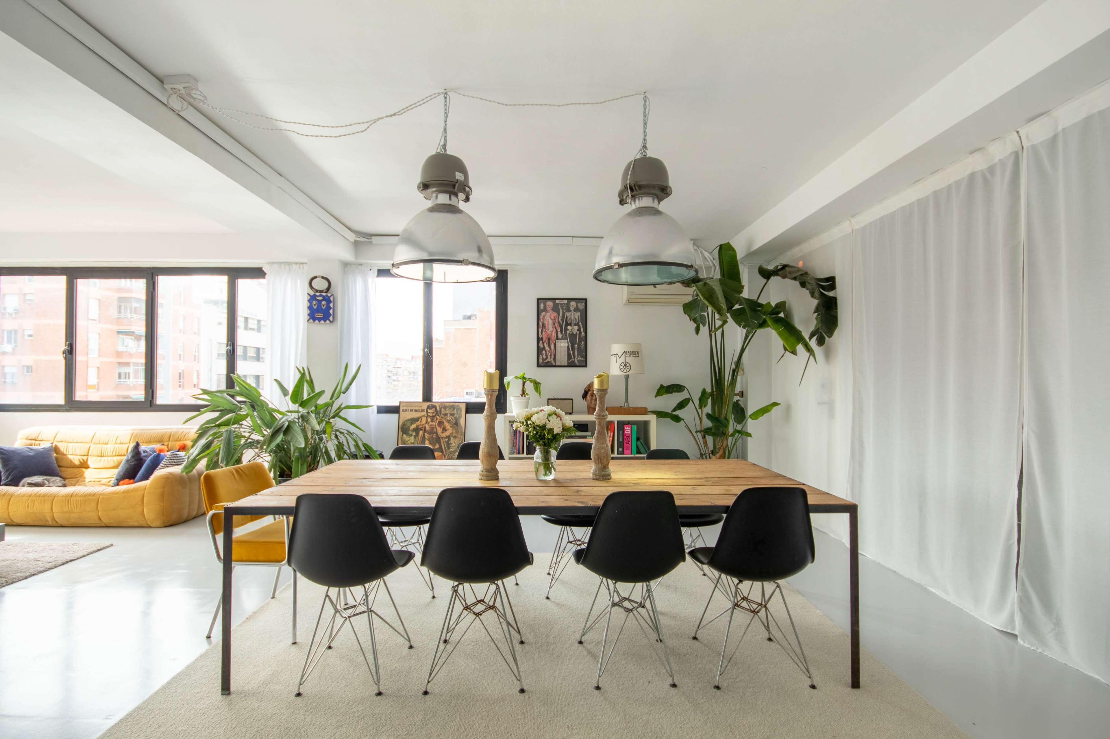 A modern dining area with a long wooden table surrounded by seven black chairs, large pendant lights hanging above, and large windows letting in natural light.