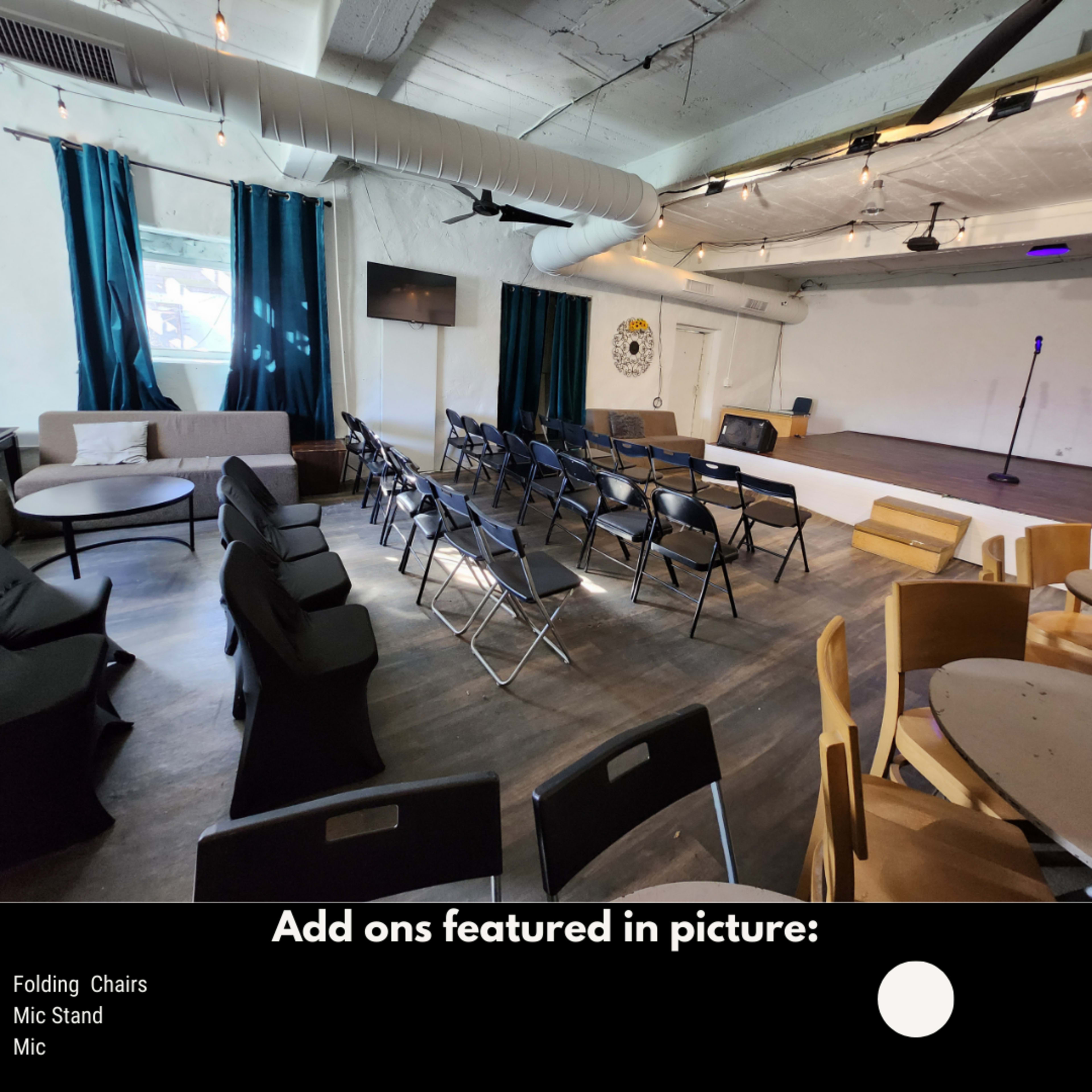 The image shows a performance space with rows of folding chairs facing a small stage equipped with a mic stand and a microphone.
