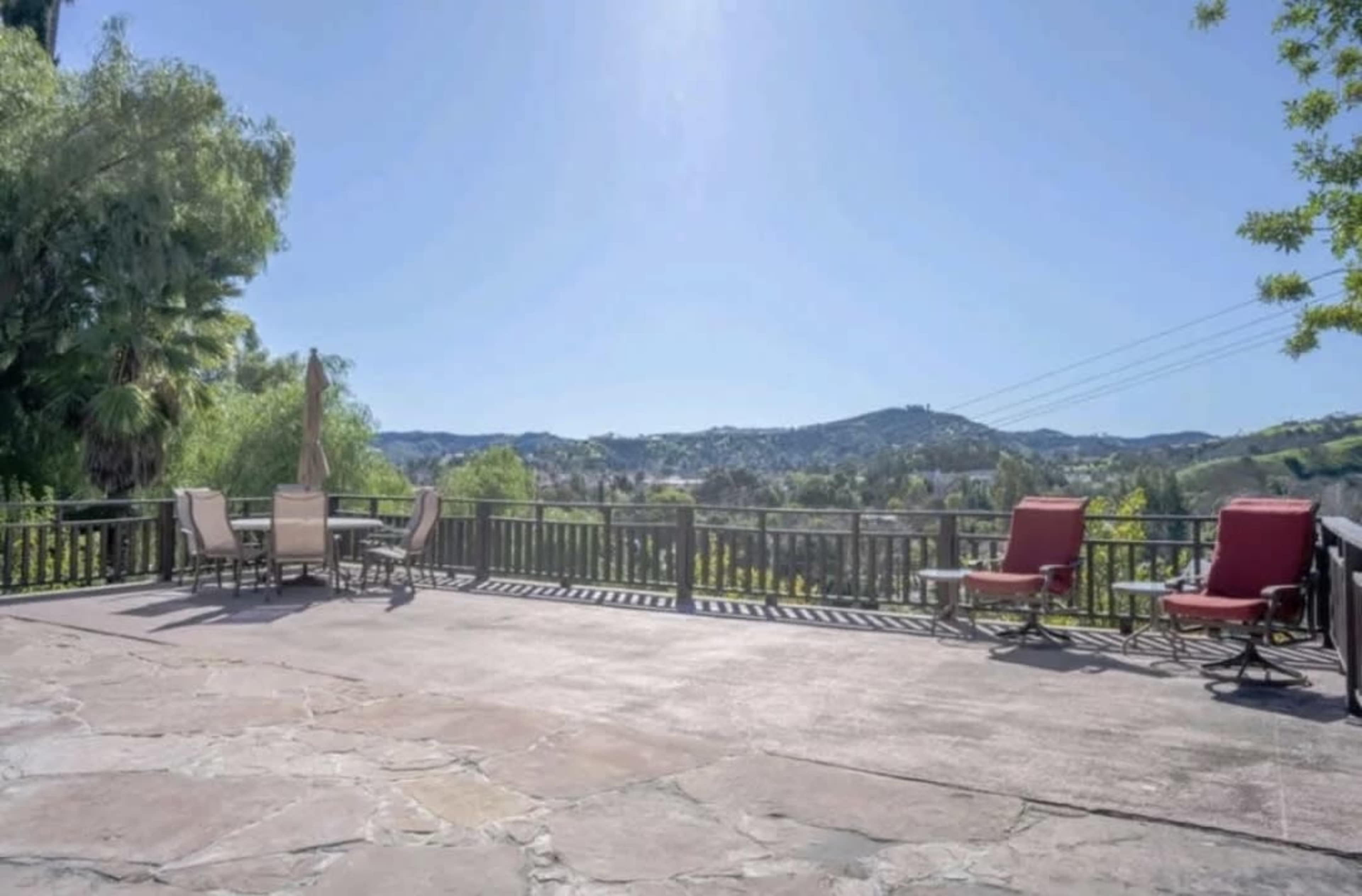 The image shows a spacious outdoor deck with seating, overlooking a mountainous landscape under a clear blue sky.