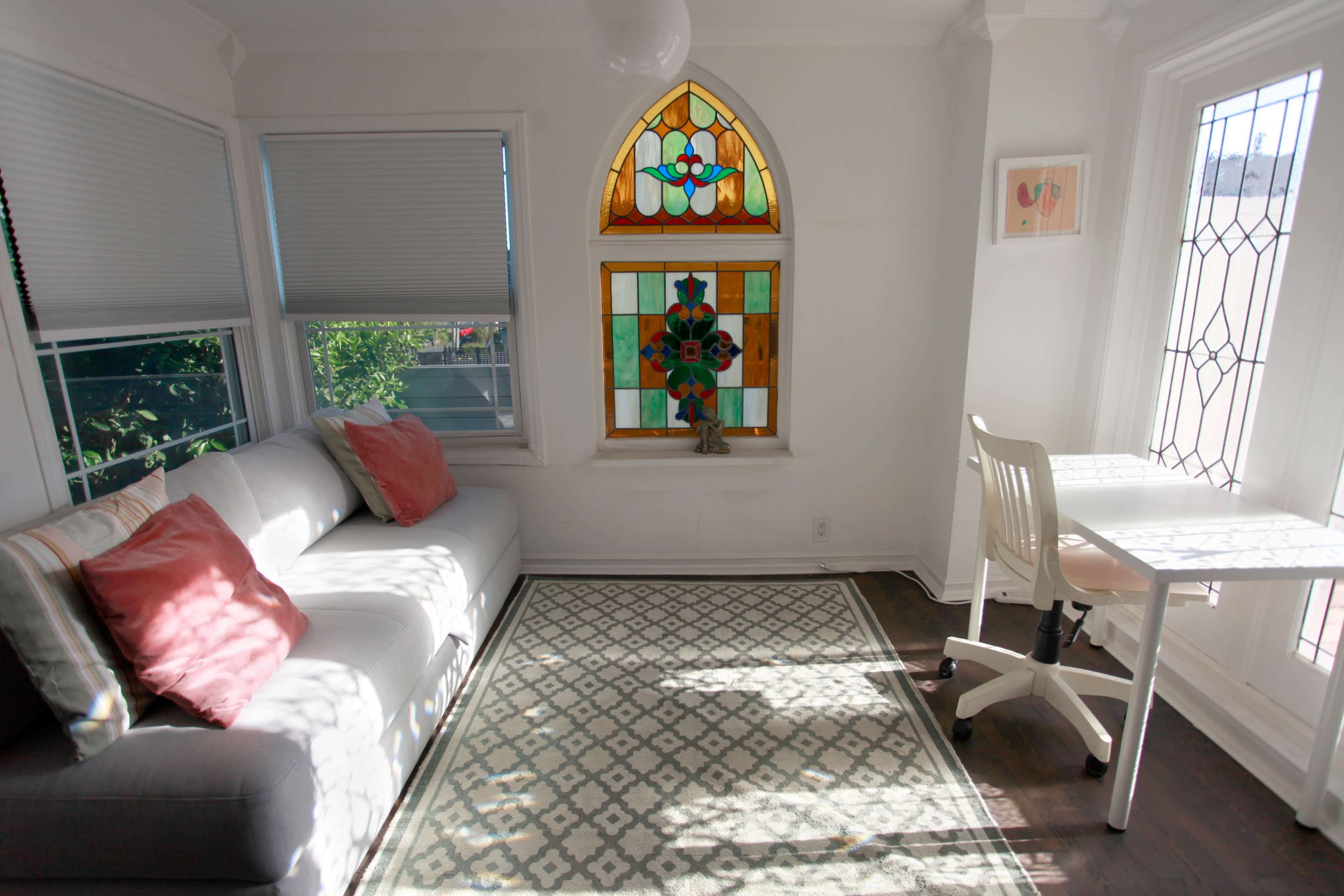 A bright room features a gray sofa with pink pillows, a stained glass window, and a white desk beside a patterned rug.