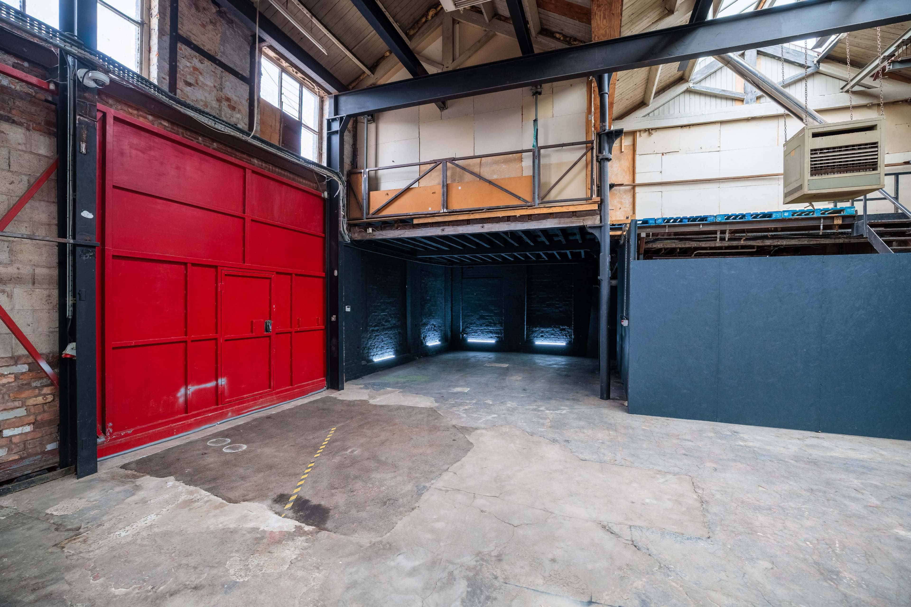 The image shows an industrial space with a red door, exposed brick walls, and a concrete floor, featuring a large open area and a raised platform.