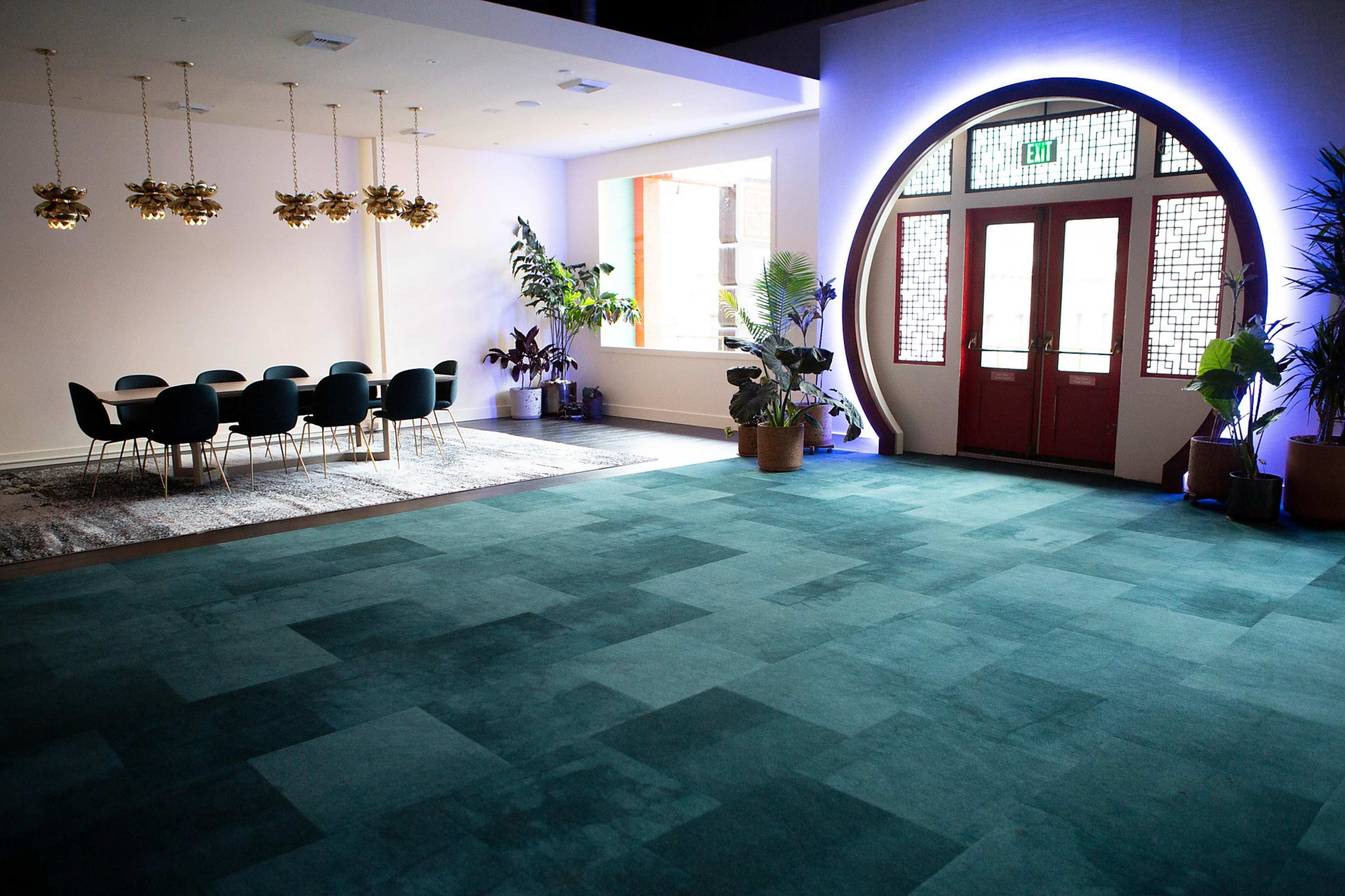 A spacious room with a teal carpet, a dining table set for eight, and large windows, along with several houseplants and decorative light fixtures.