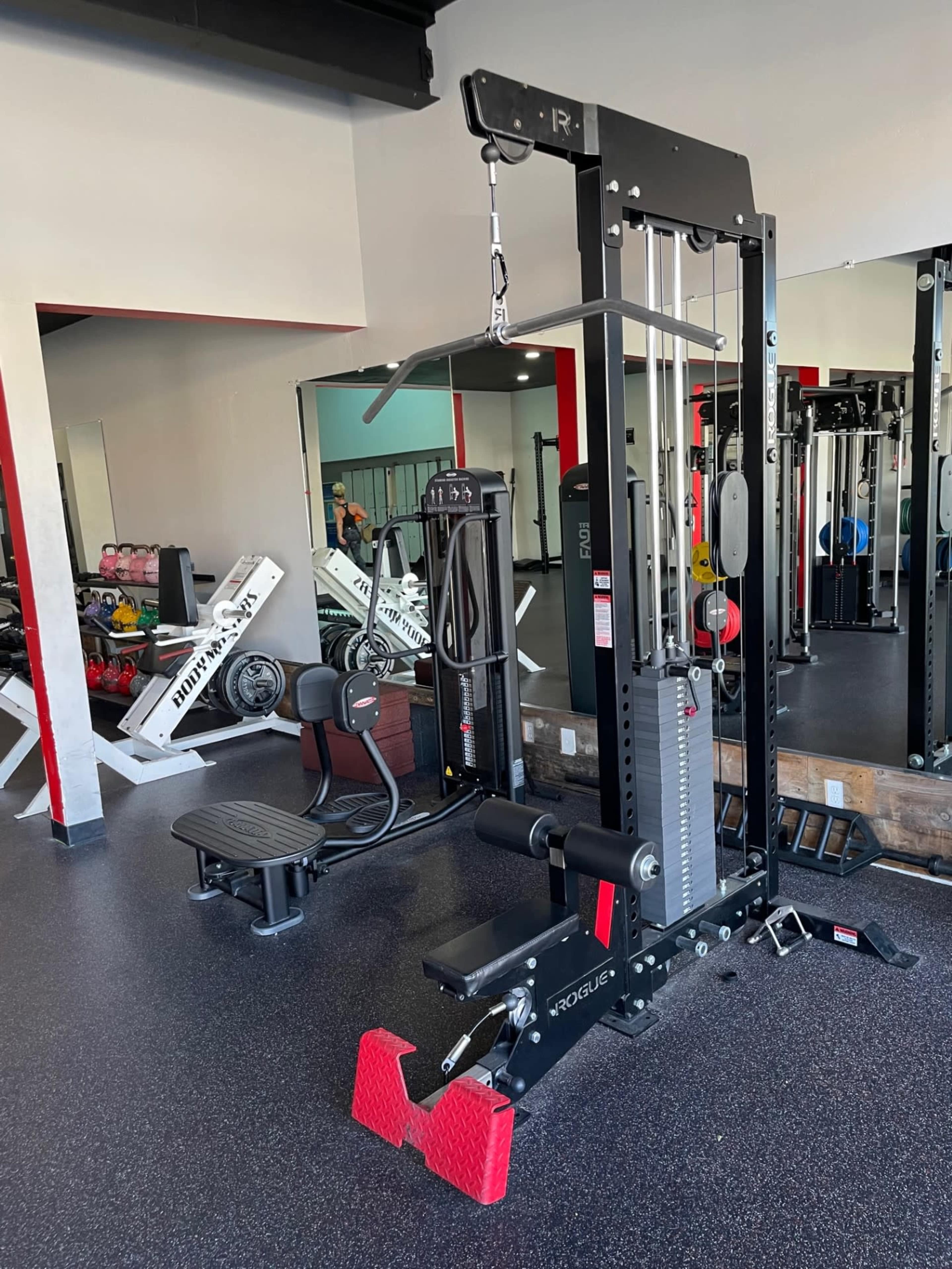 A gym interior featuring a cable machine with various weights and an upright bench nearby, surrounded by exercise equipment.