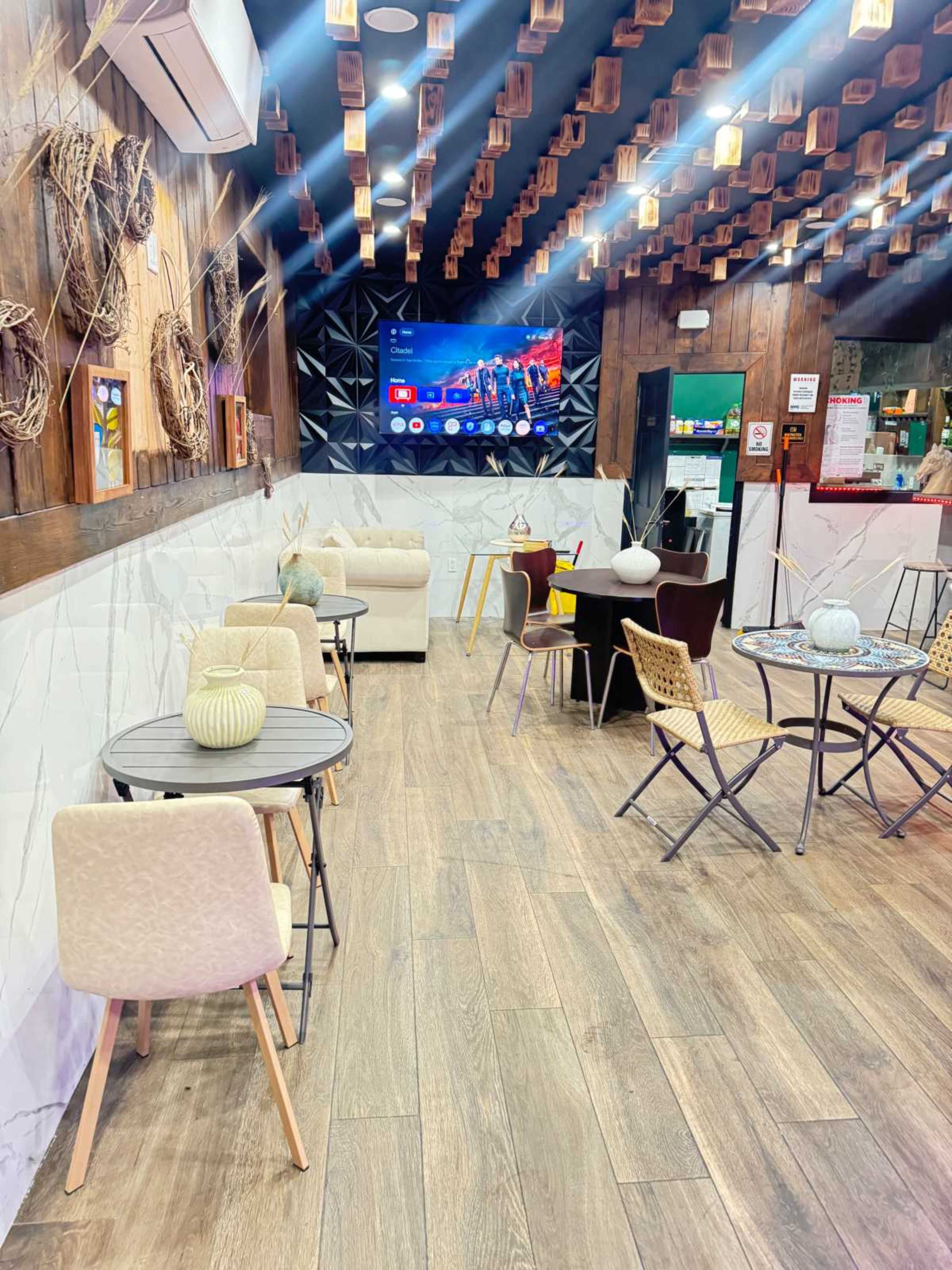 The image depicts a modern café interior with seating arrangements including small tables and chairs, a large television screen on a feature wall, and decorative elements overhead.