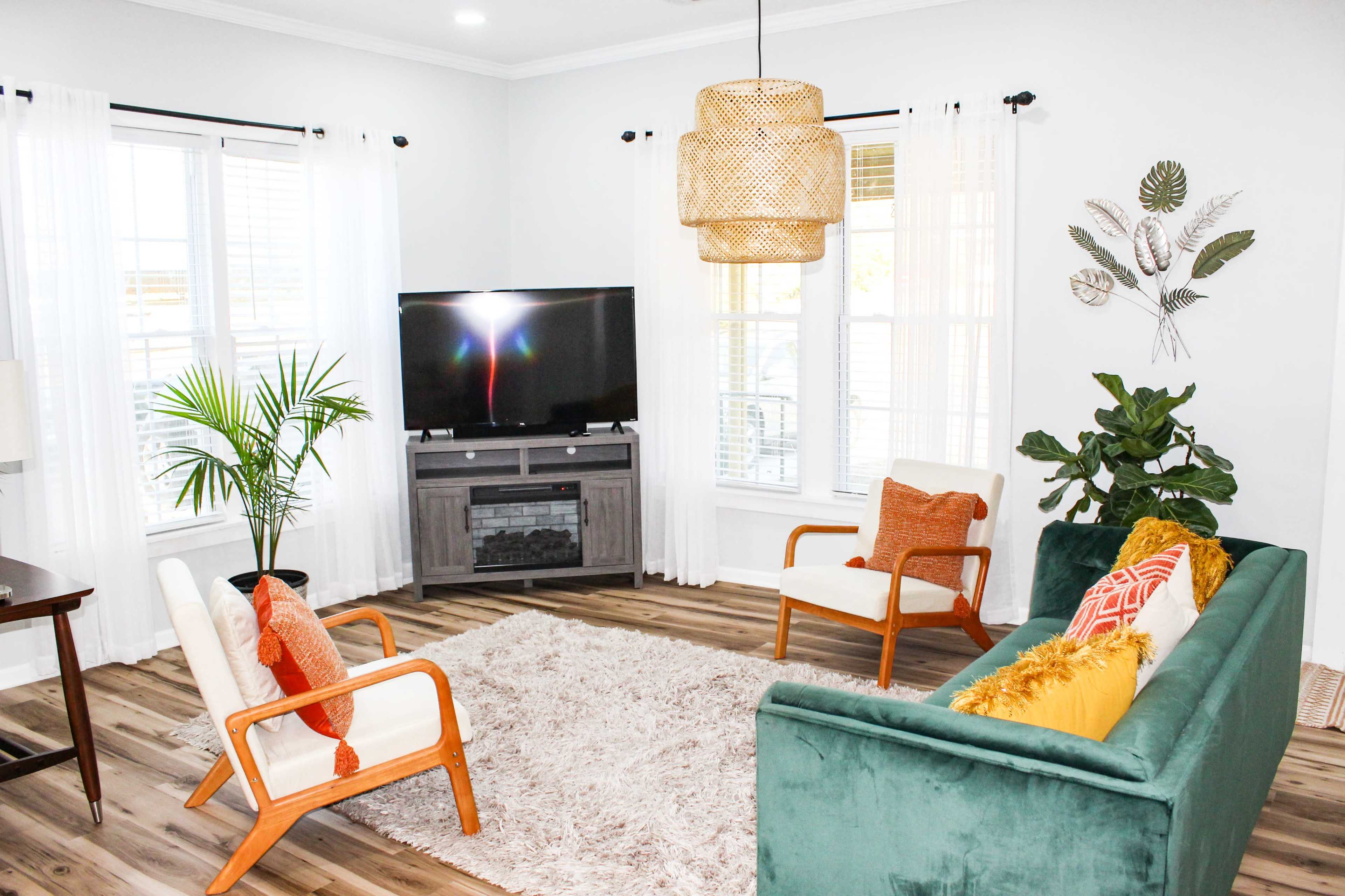 A modern living room features a green sofa, two wooden armchairs, a large area rug, and a television on a cabinet, with bright natural light coming through the windows.