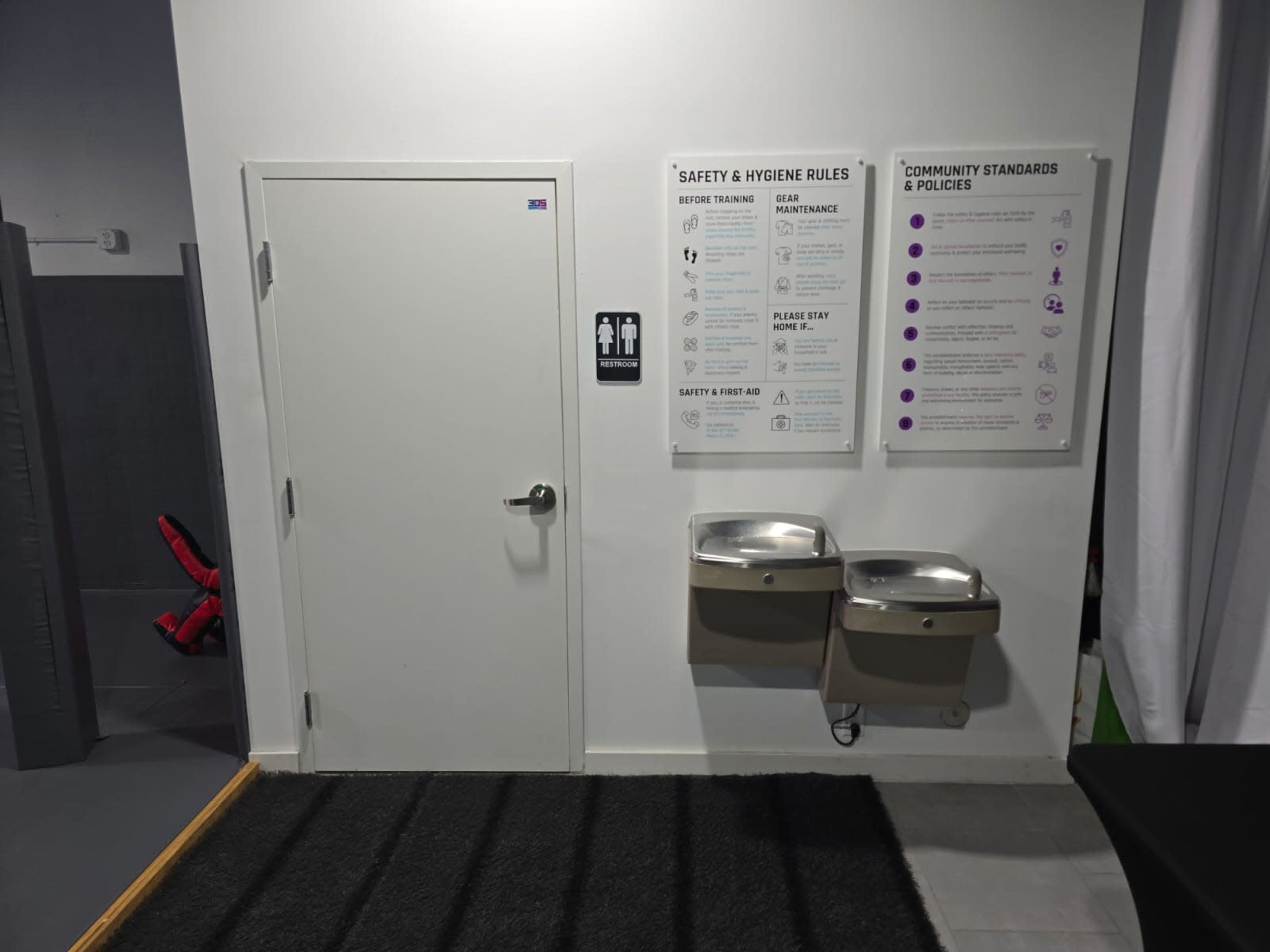 The image shows a door labeled with a restroom sign next to two drinking fountains and a wall displaying safety and hygiene rules.