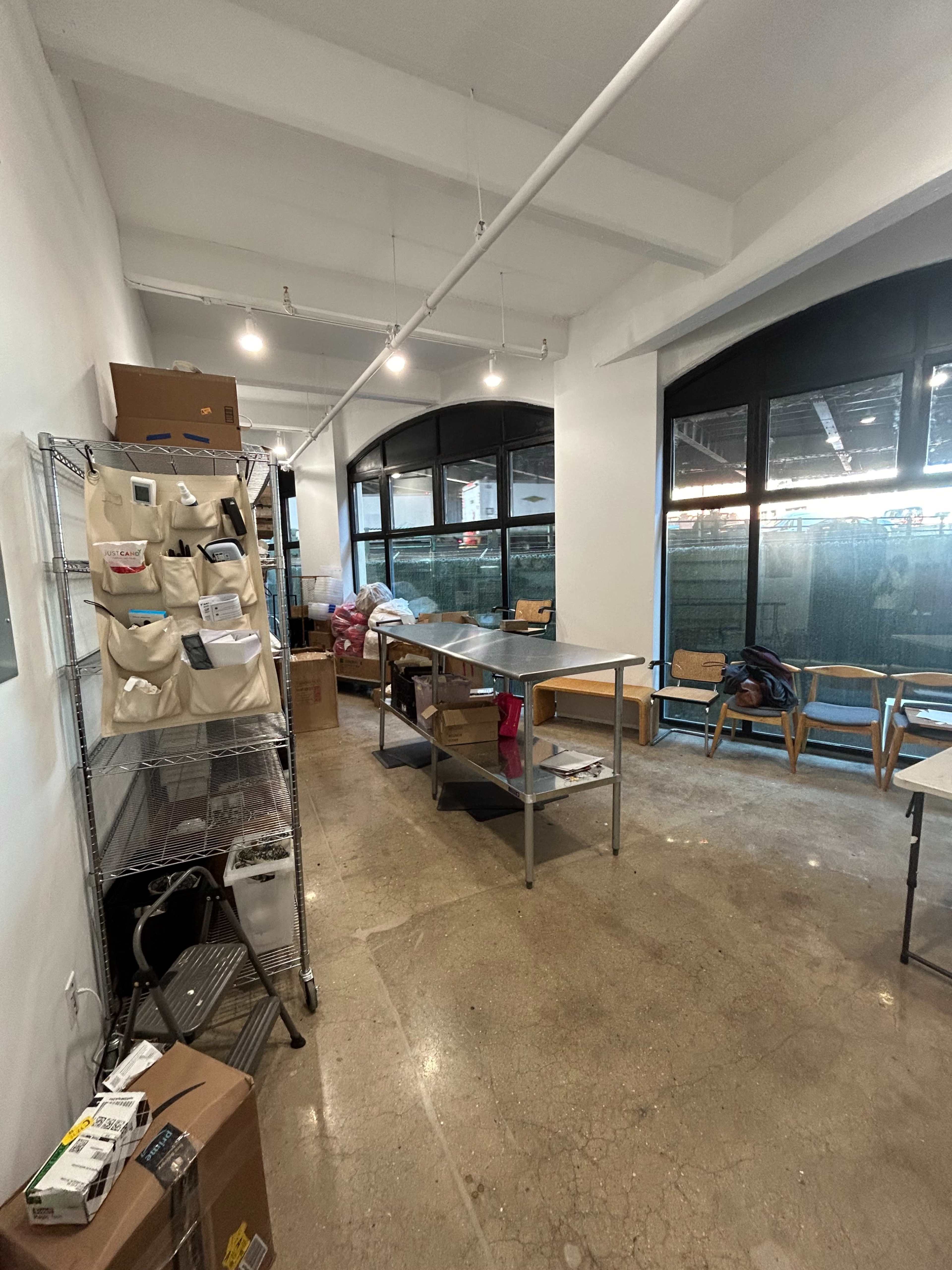 The image shows a large, empty room with a concrete floor, tables in the center, shelves with storage bags on one side, and large windows letting in natural light.