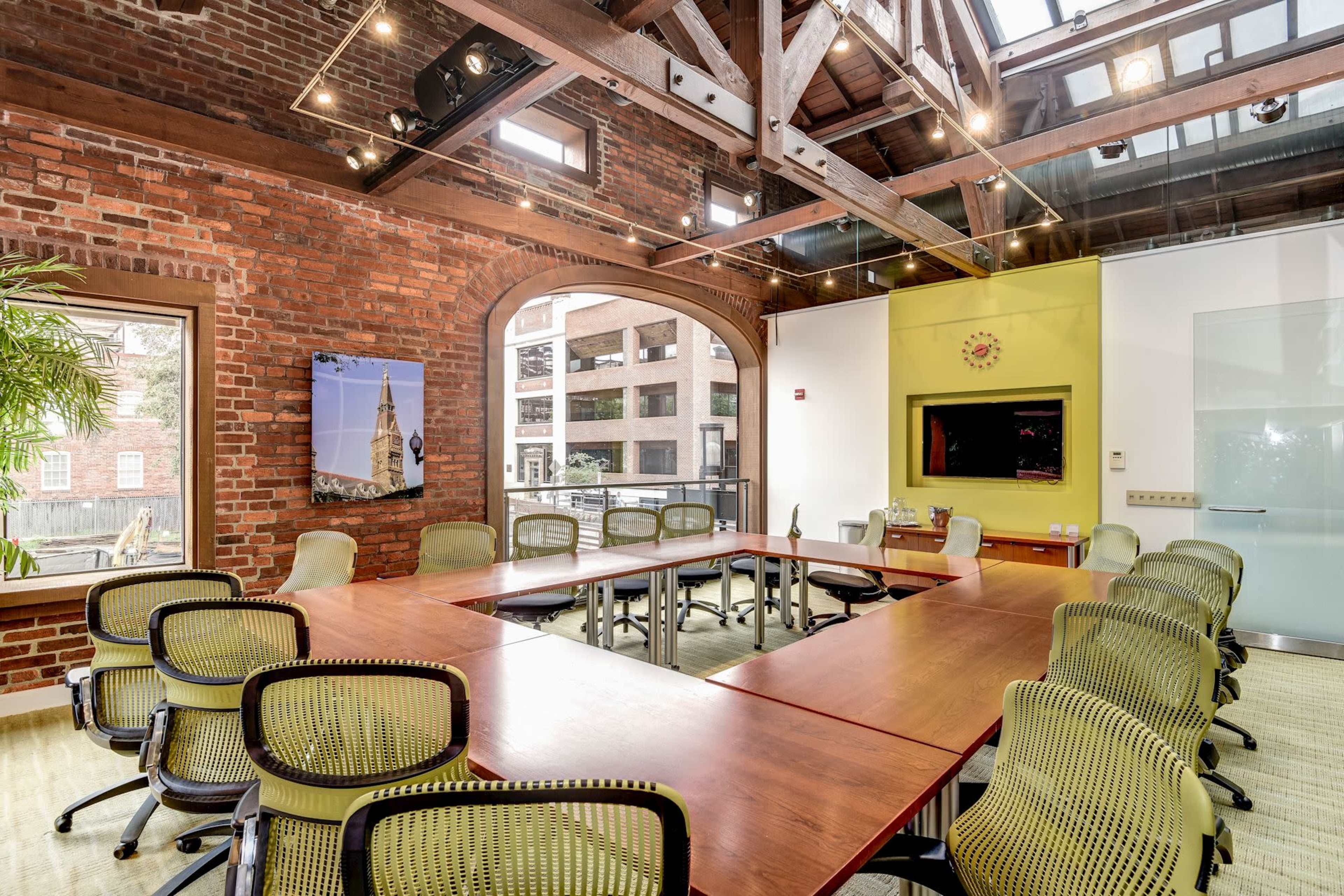 A modern conference room features a long wooden table surrounded by green chairs, with exposed brick walls and large windows allowing natural light.