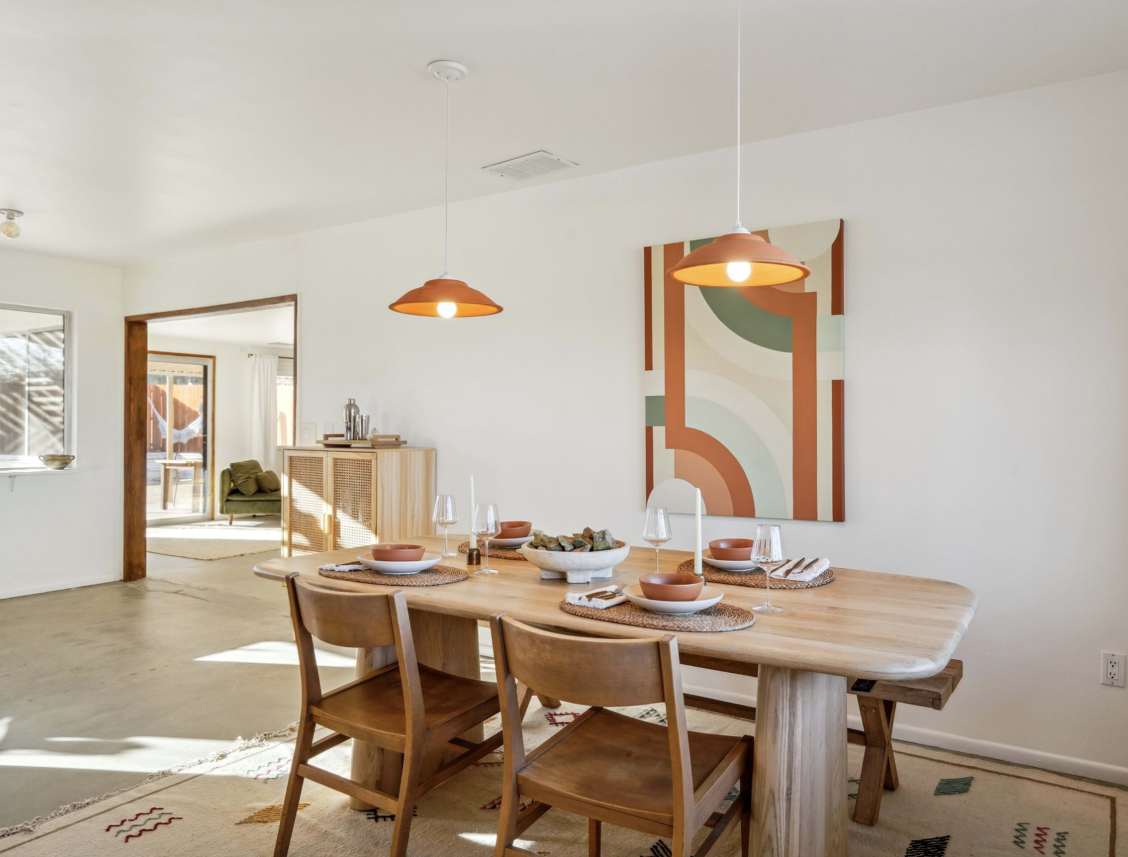 A dining area features a wooden table with chairs, pendant lighting, and an abstract wall art piece, with a view toward a living space.