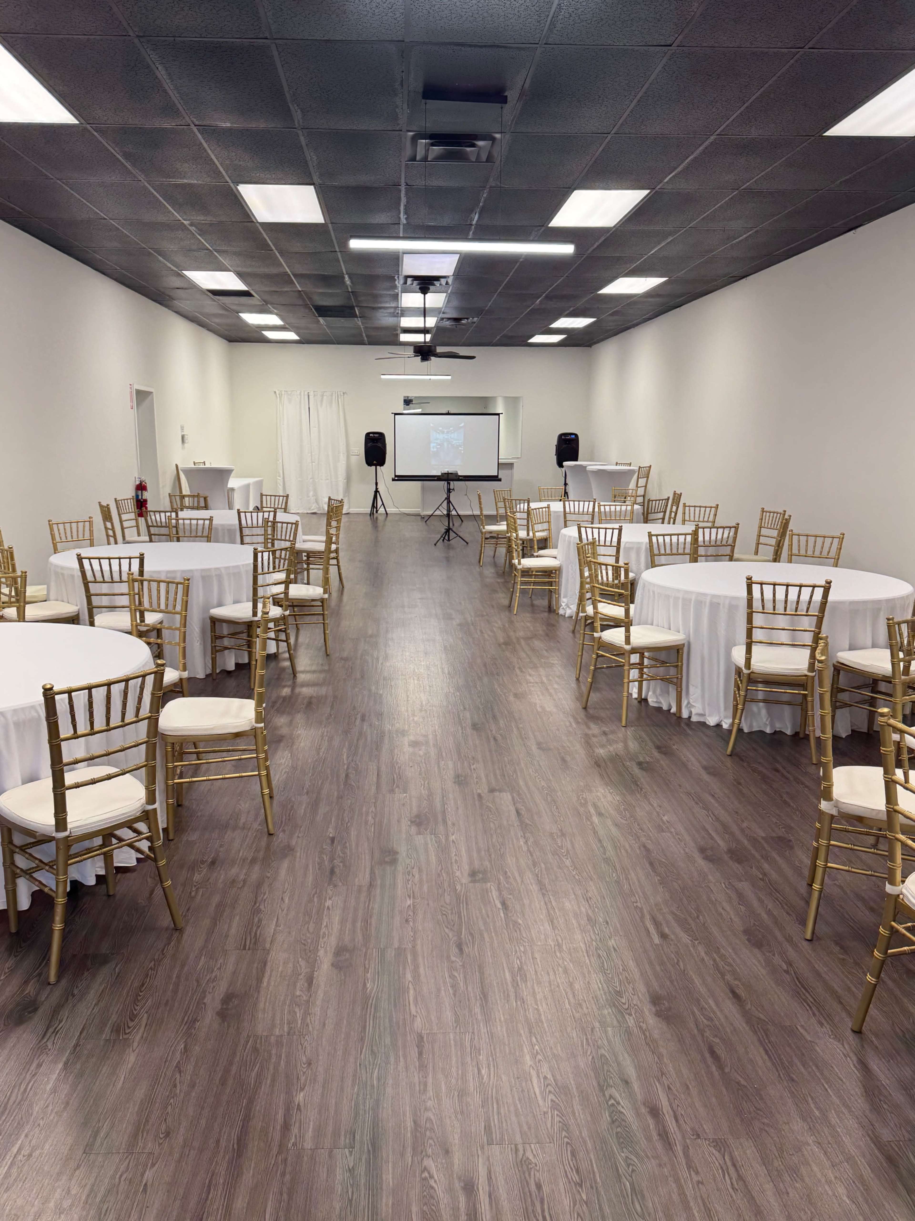 The image shows a spacious event room with round tables covered in white linens and gold chairs arranged neatly, and a presentation screen at the far end.