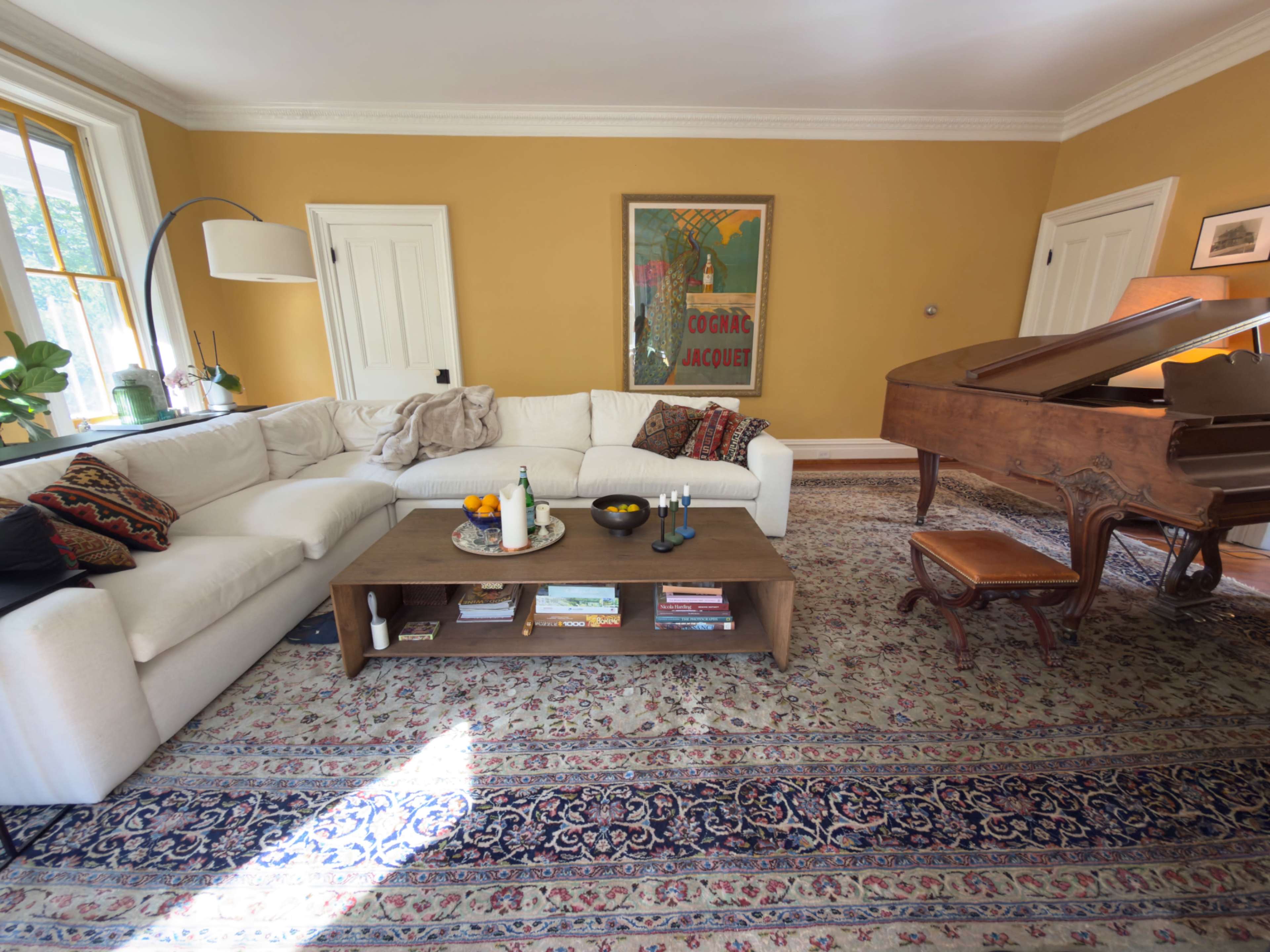 A spacious living room features a white sectional sofa, a wooden coffee table with decorative items, and a grand piano against a mustard-yellow wall.