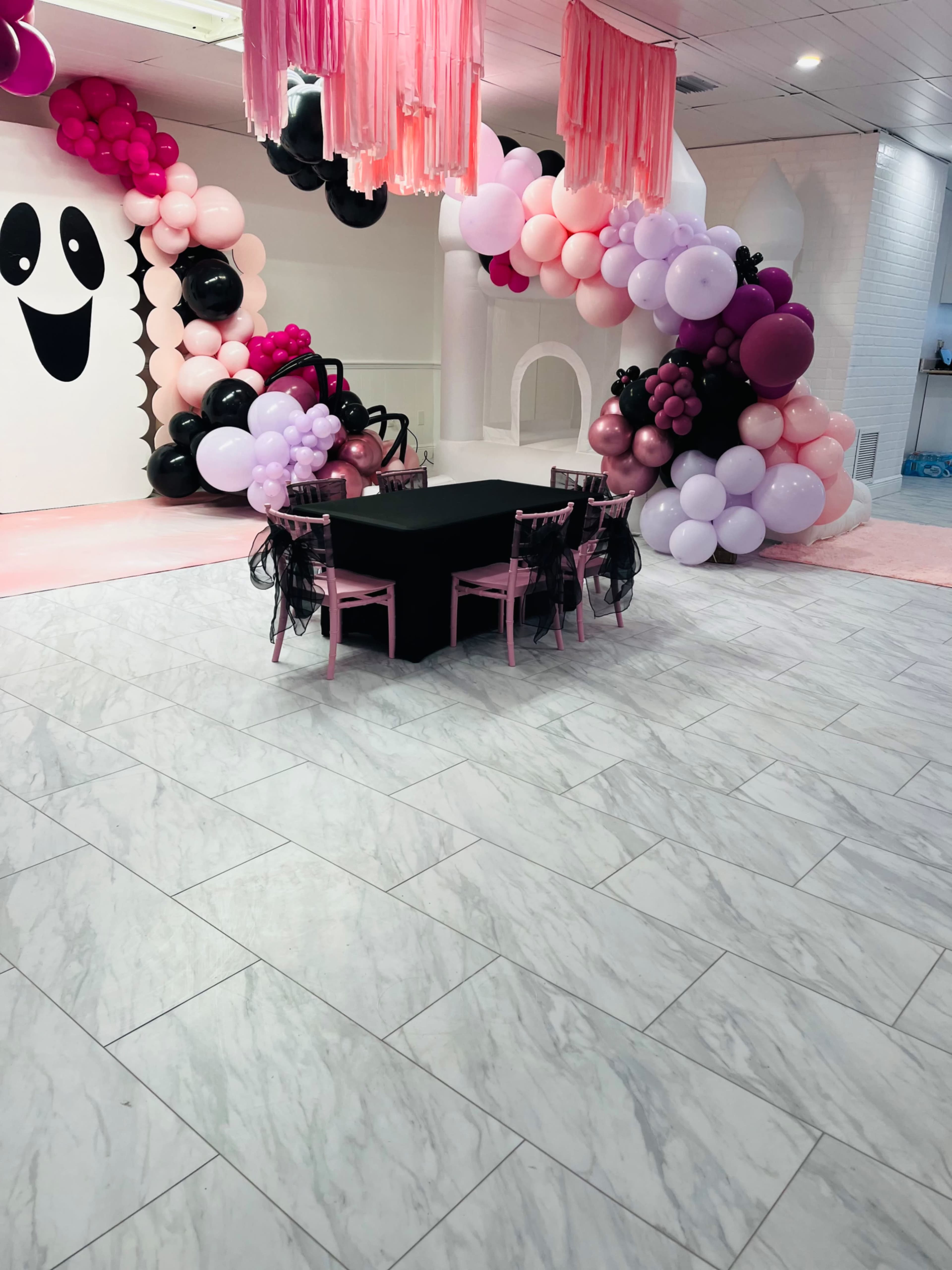 A decorated event space featuring a black table with pink chairs surrounded by colorful balloon arrangements and a wall with a ghost design.