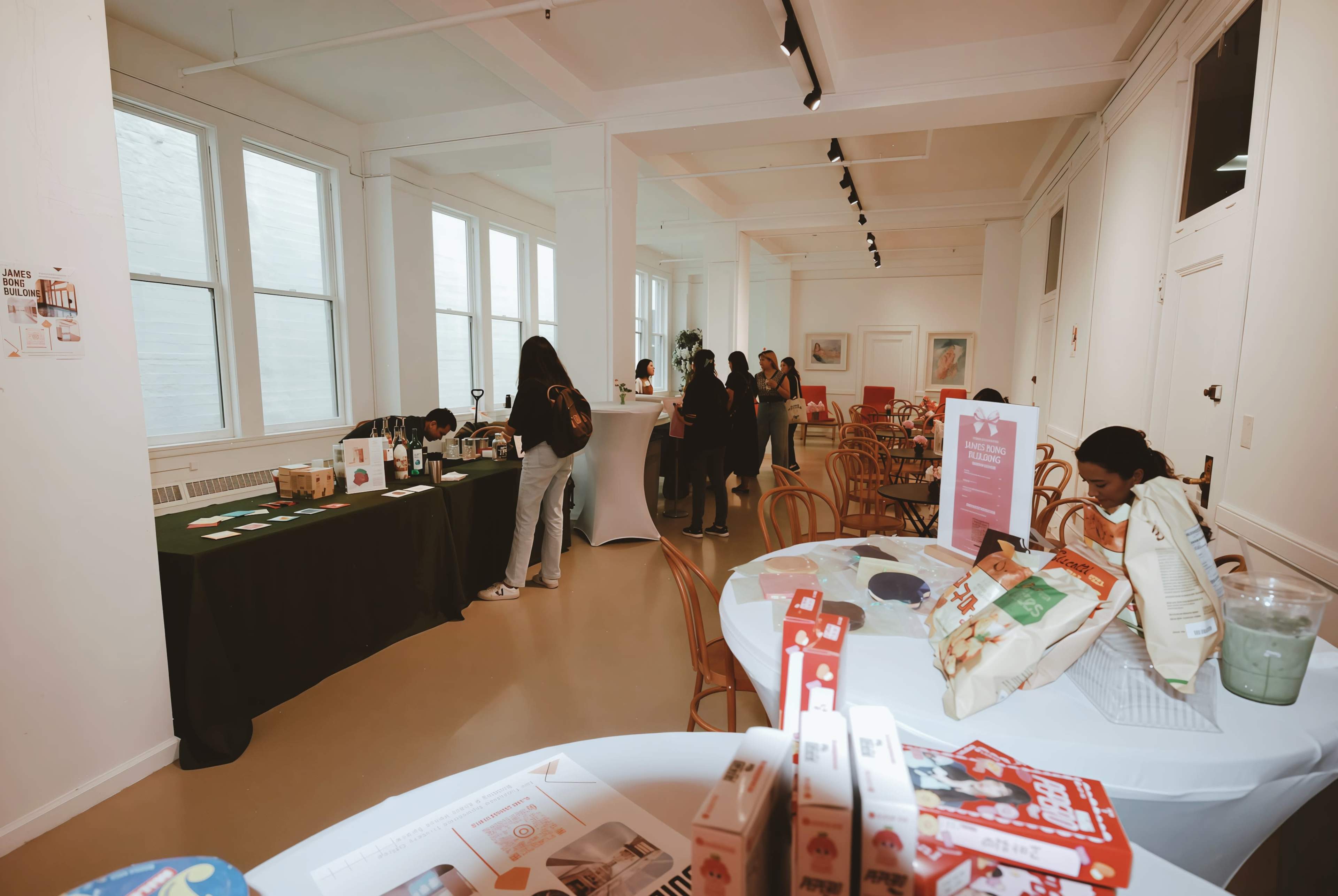 The image shows a spacious, well-lit room with tables set up for an event, featuring food and beverage displays on one side and people gathered in conversation on the other.