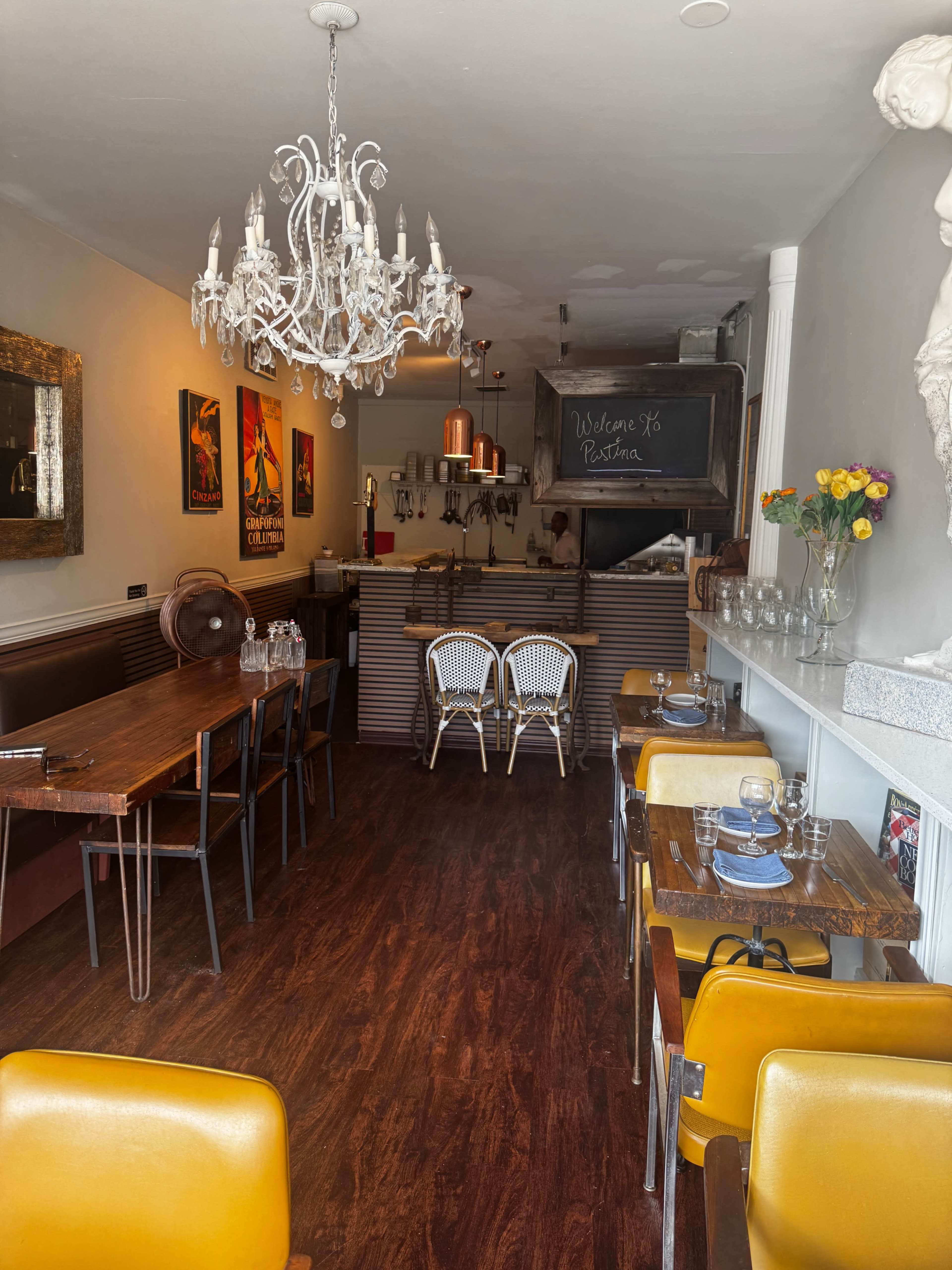 The image shows a cozy restaurant interior featuring wooden tables, yellow chairs, a chandelier, and a chalkboard sign welcoming guests.