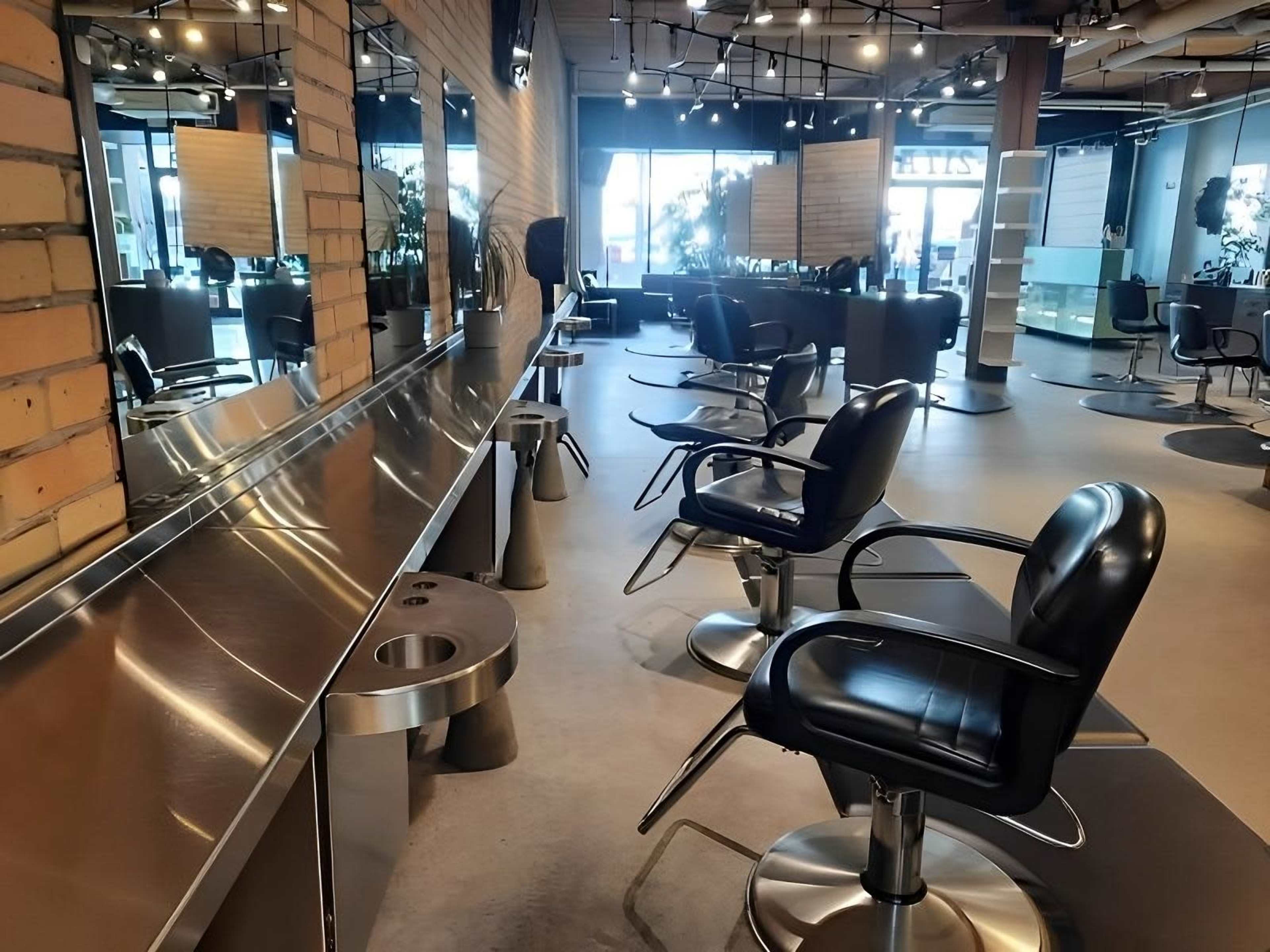A modern hair salon featuring several black salon chairs positioned along a stainless steel counter, with large mirrors and a bright, spacious layout.