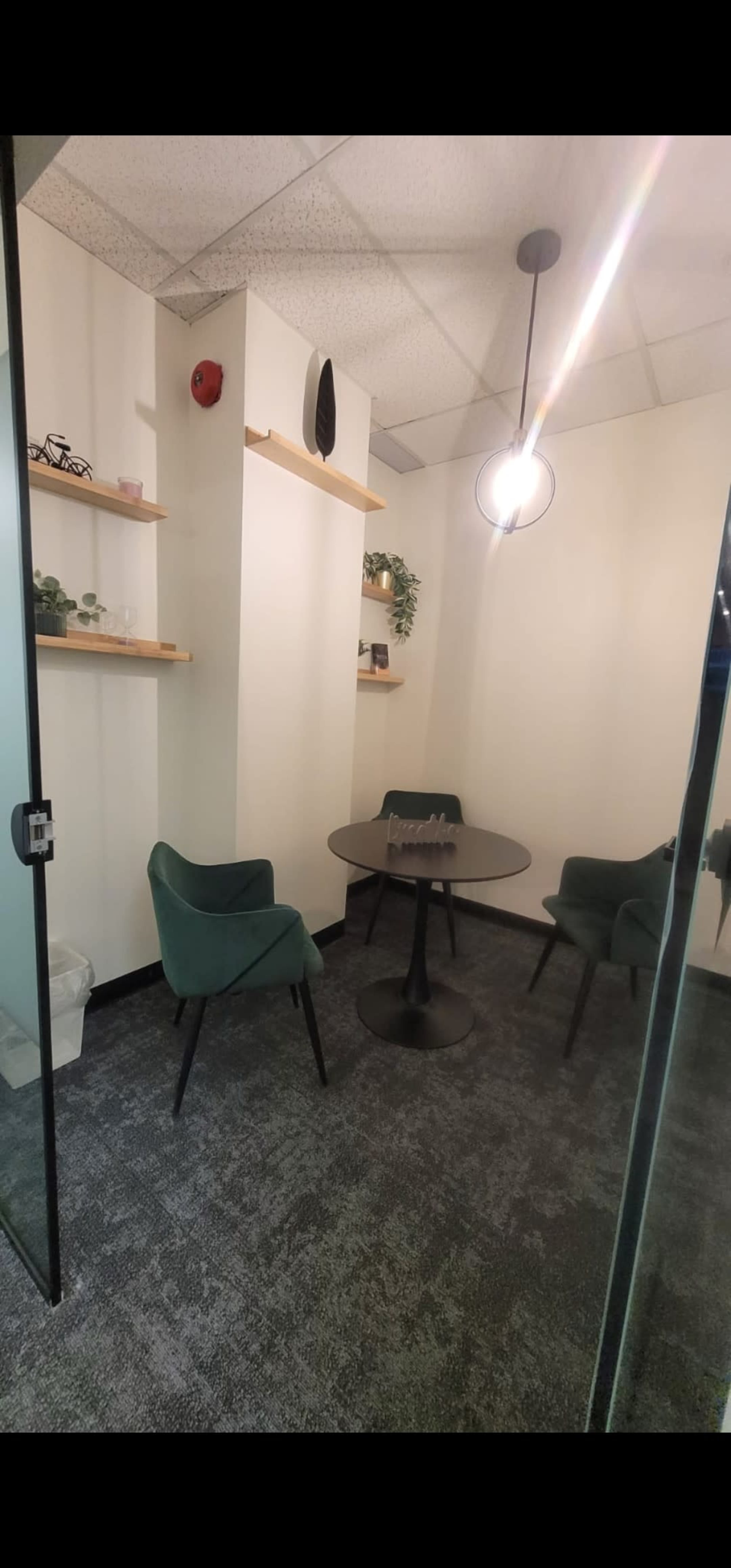 The image shows a small office meeting room with a round table and four green chairs, featuring shelves with decorative items and plants on the wall.