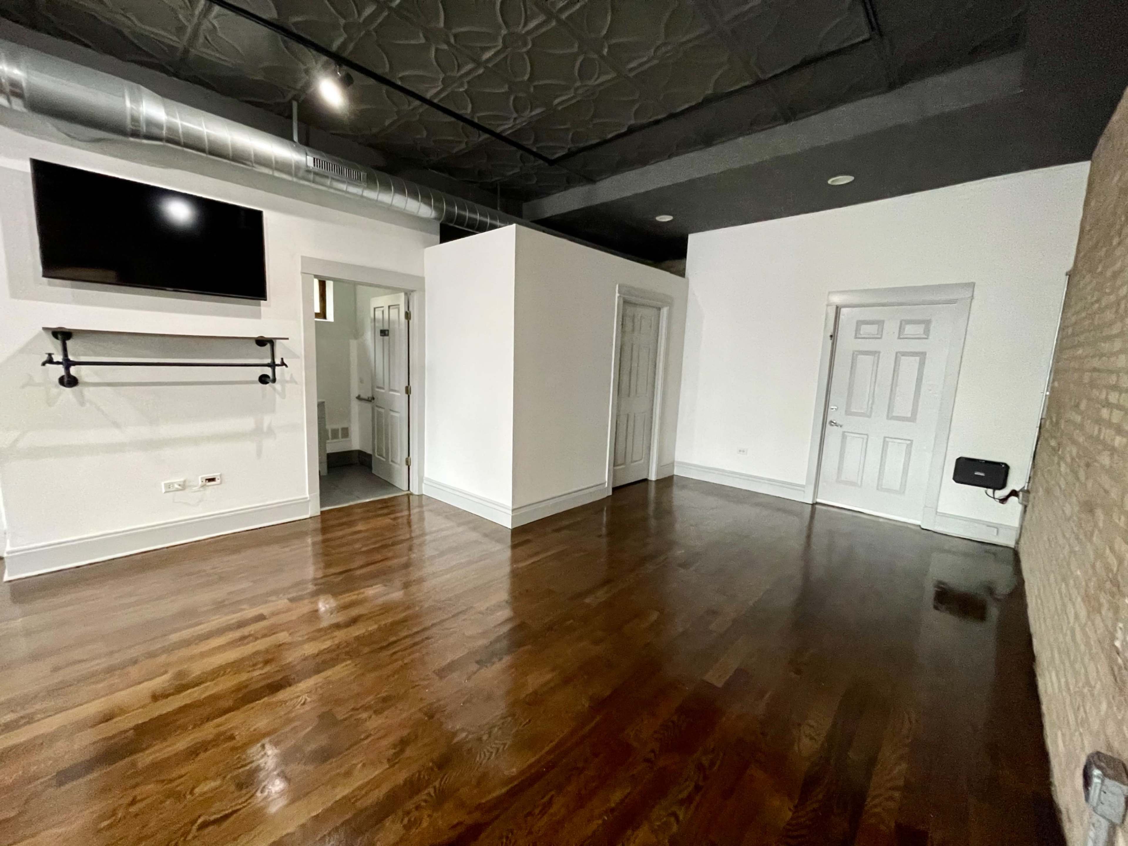 A spacious room with hardwood floors, a flat-screen TV mounted on the wall, and various doorways leading to other areas.