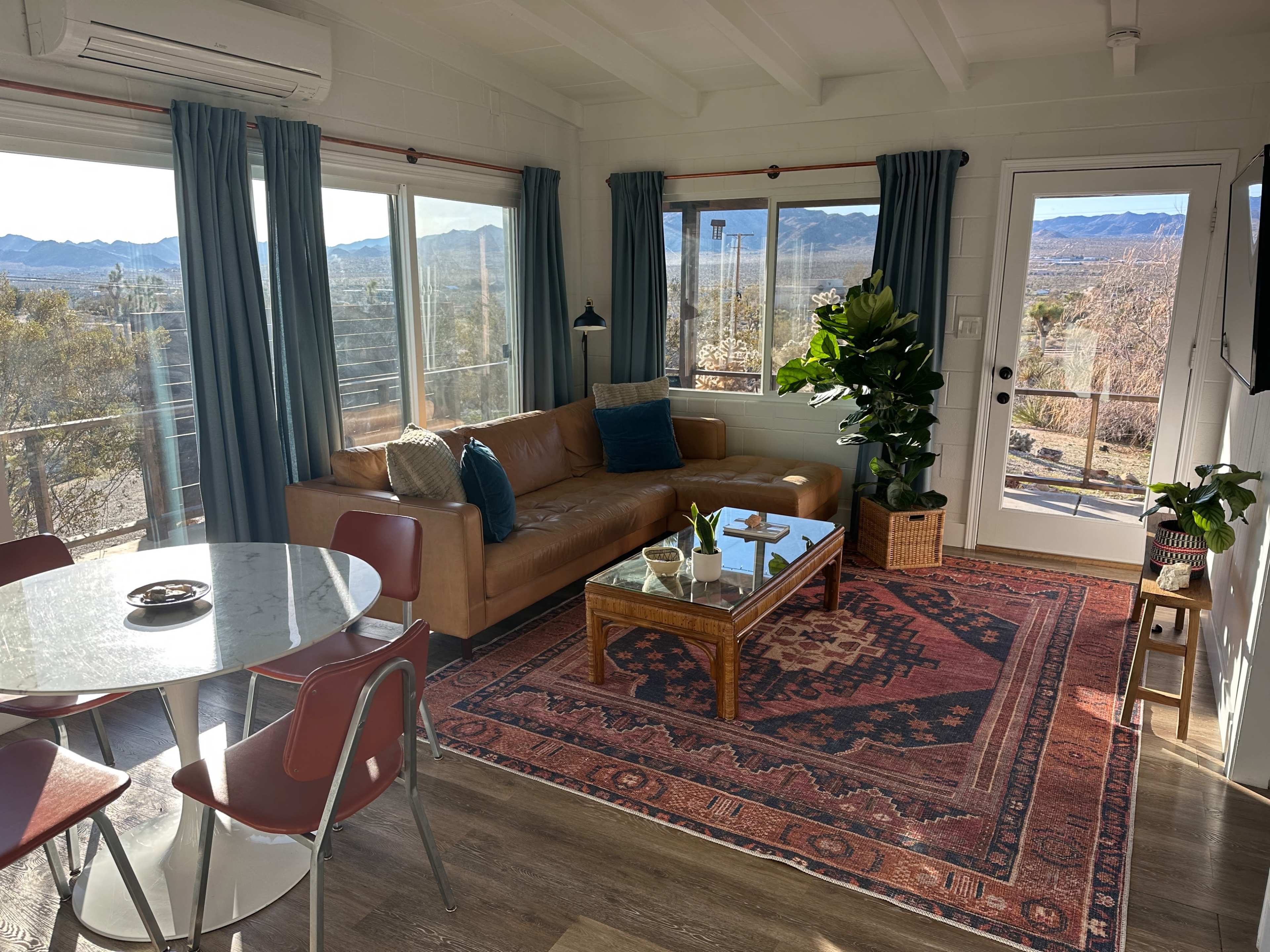 A living area featuring a brown leather couch, a glass coffee table on a patterned rug, a dining table with red chairs, large windows with mountain views, and potted plants.