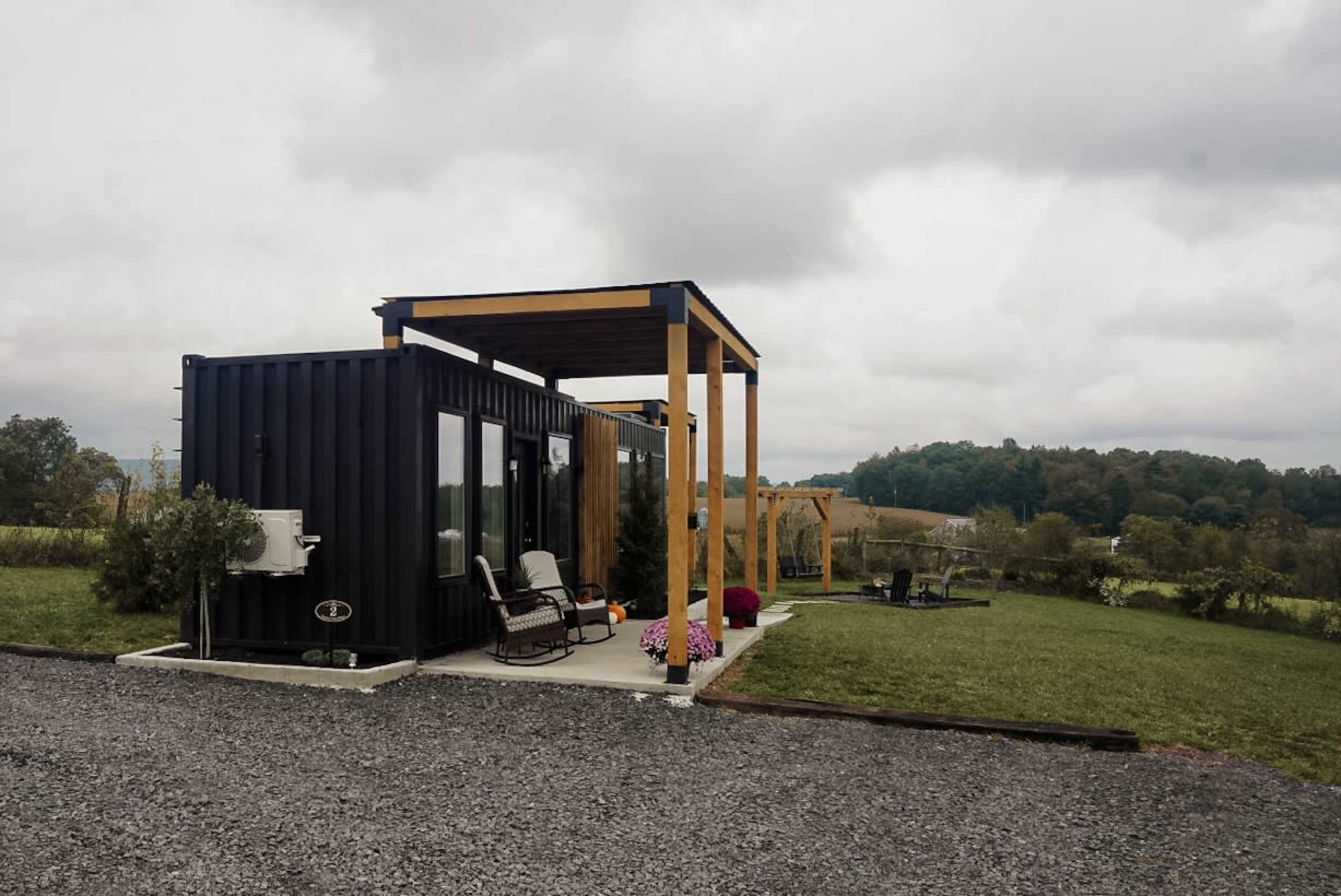 A modern black shipping container home is set on a gravel driveway with a covered porch, surrounded by a grassy landscape and distant hills.
