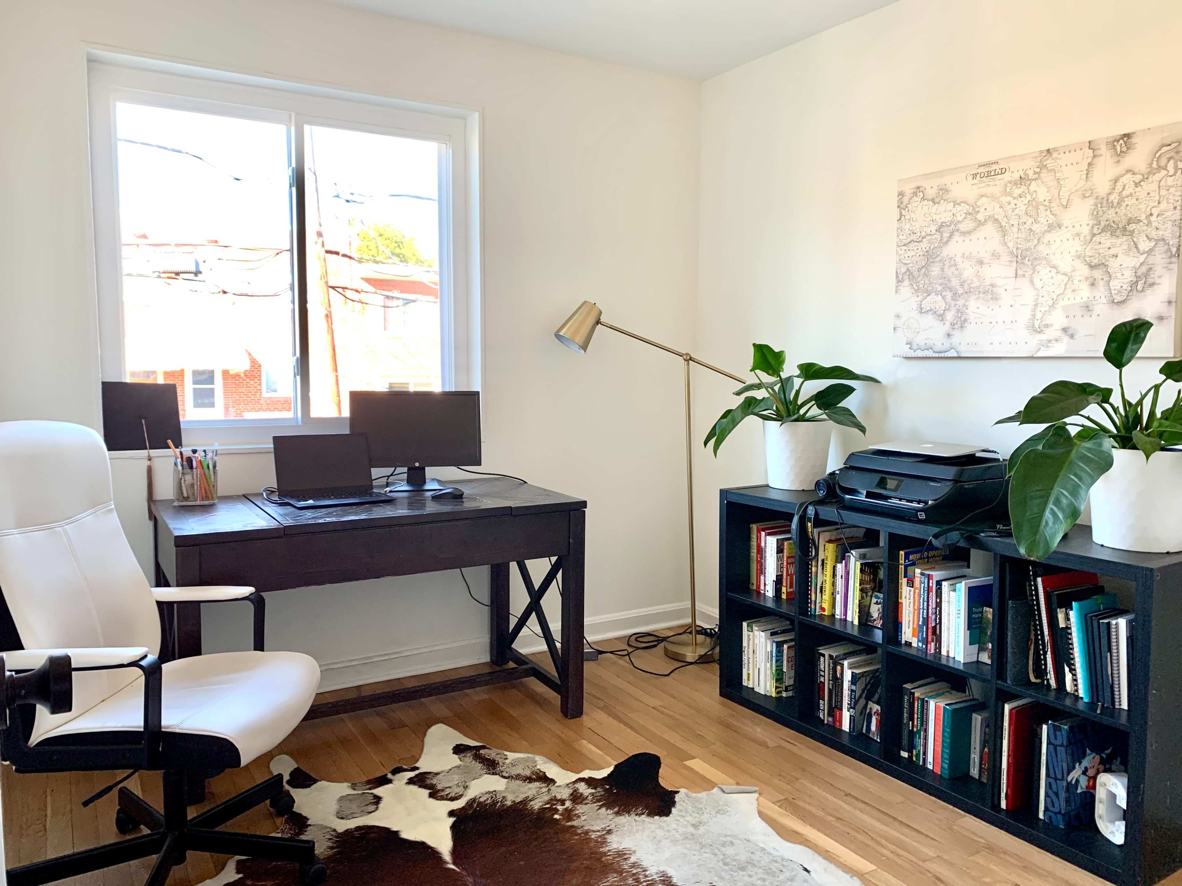 A white office features a desk with two monitors, a leather chair, a bookshelf filled with books, and plants beside a window.
