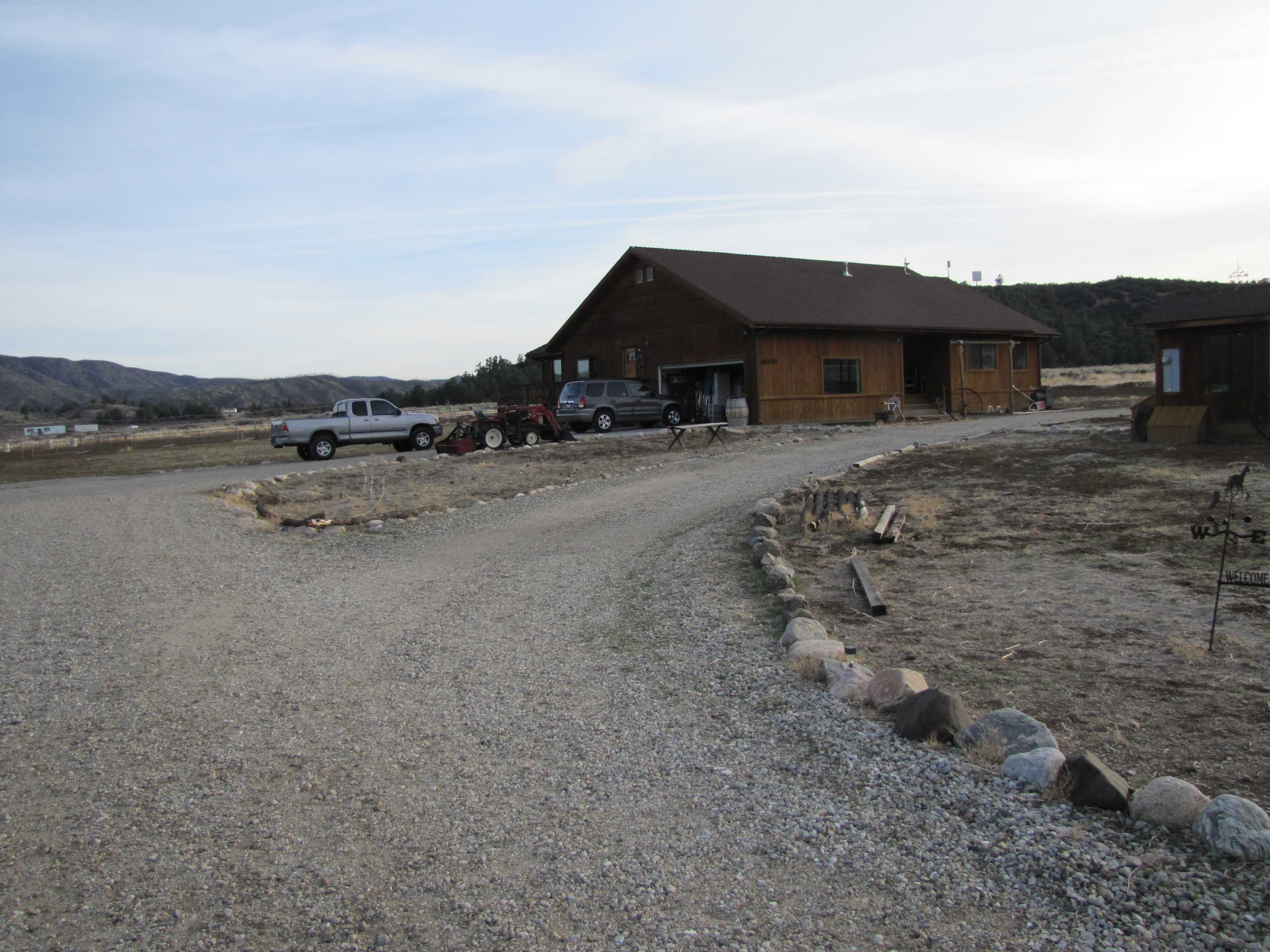 A gravel driveway leads to a large wooden house with a parking area for vehicles and a tractor in a rural setting.