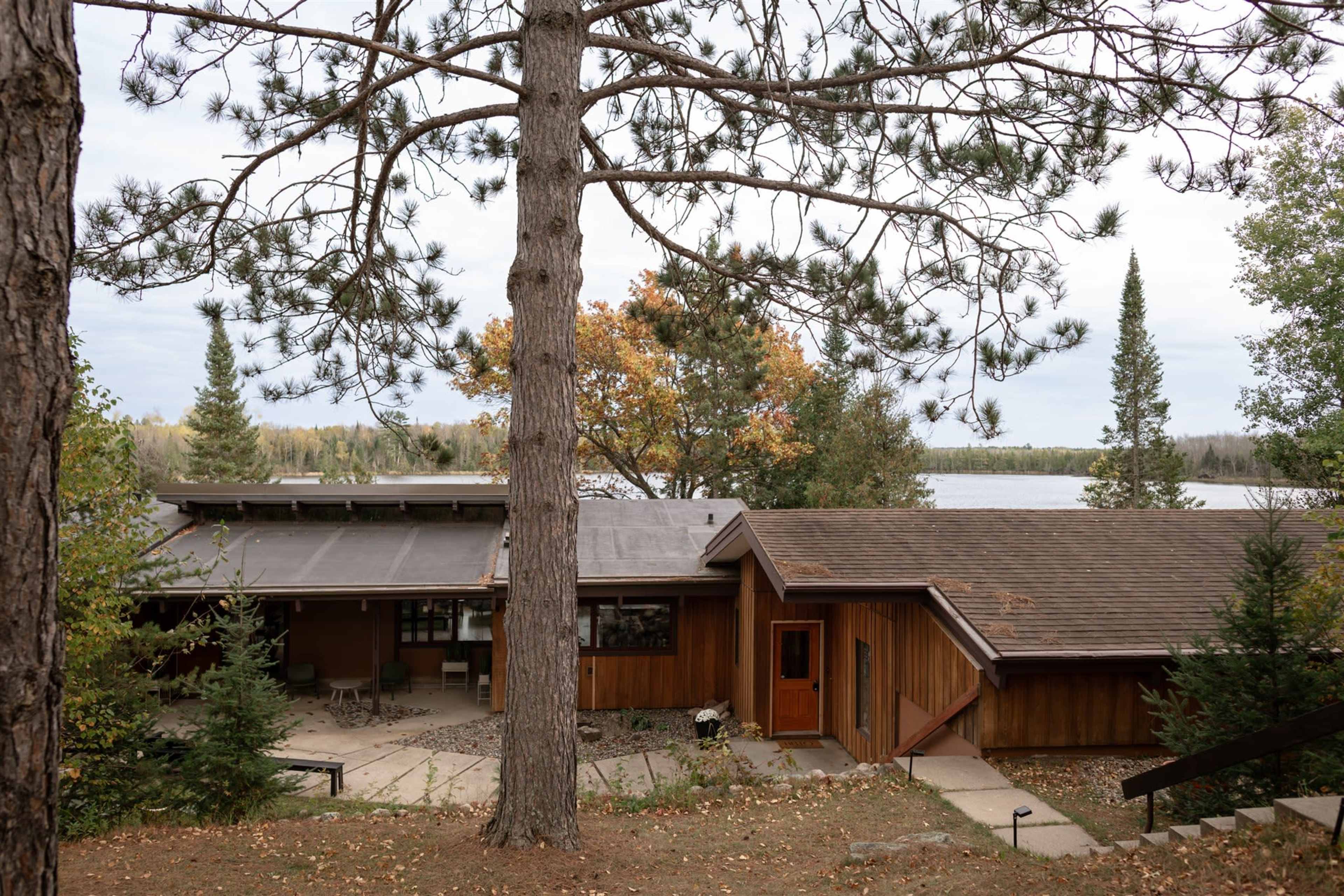 A wooden cabin is situated near a lake, surrounded by trees and autumn foliage.
