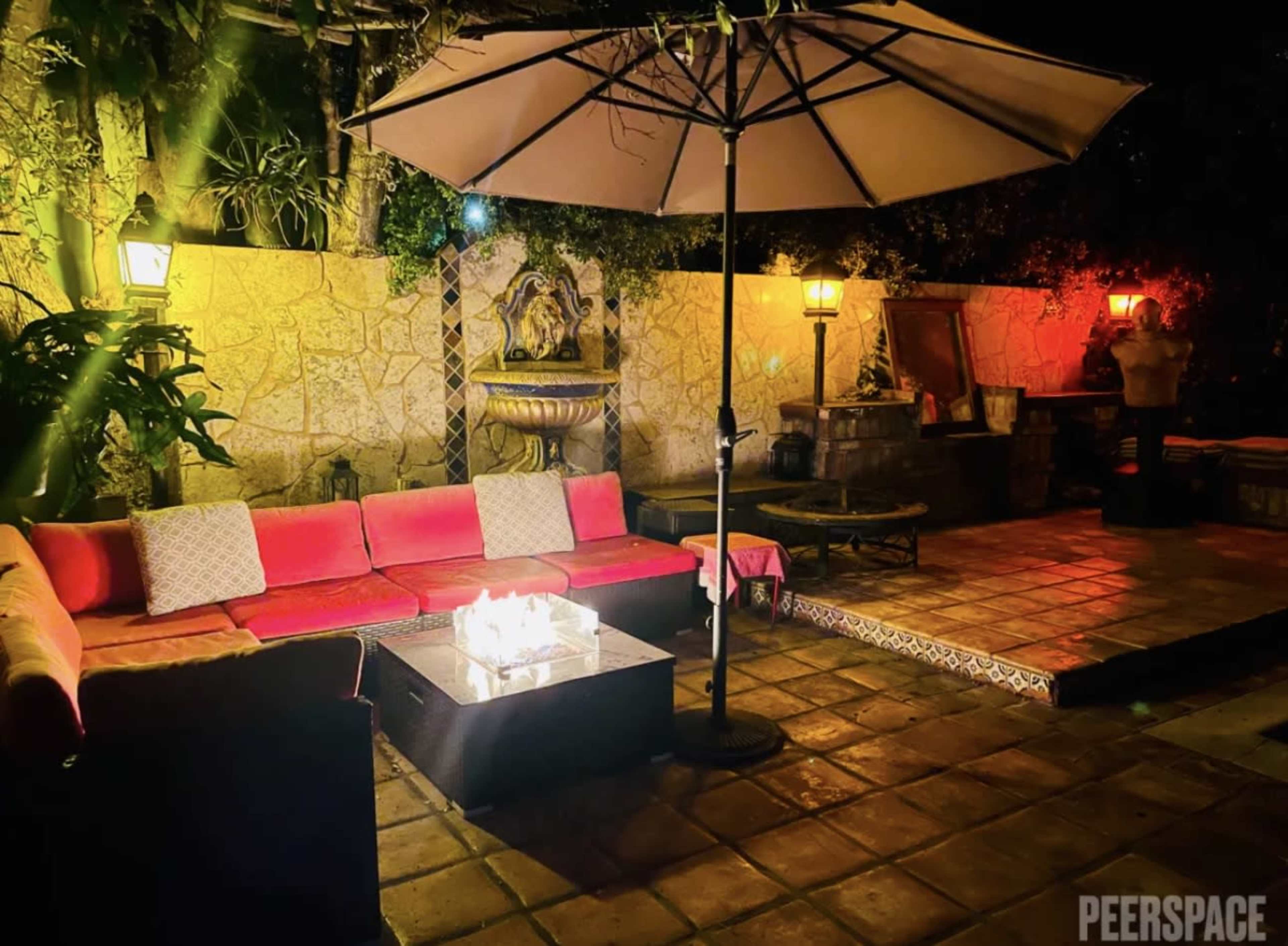 The image shows a patio area at night with a large pink sofa, a fire pit, and an umbrella over a seating arrangement, surrounded by stone walls and decorative lights.