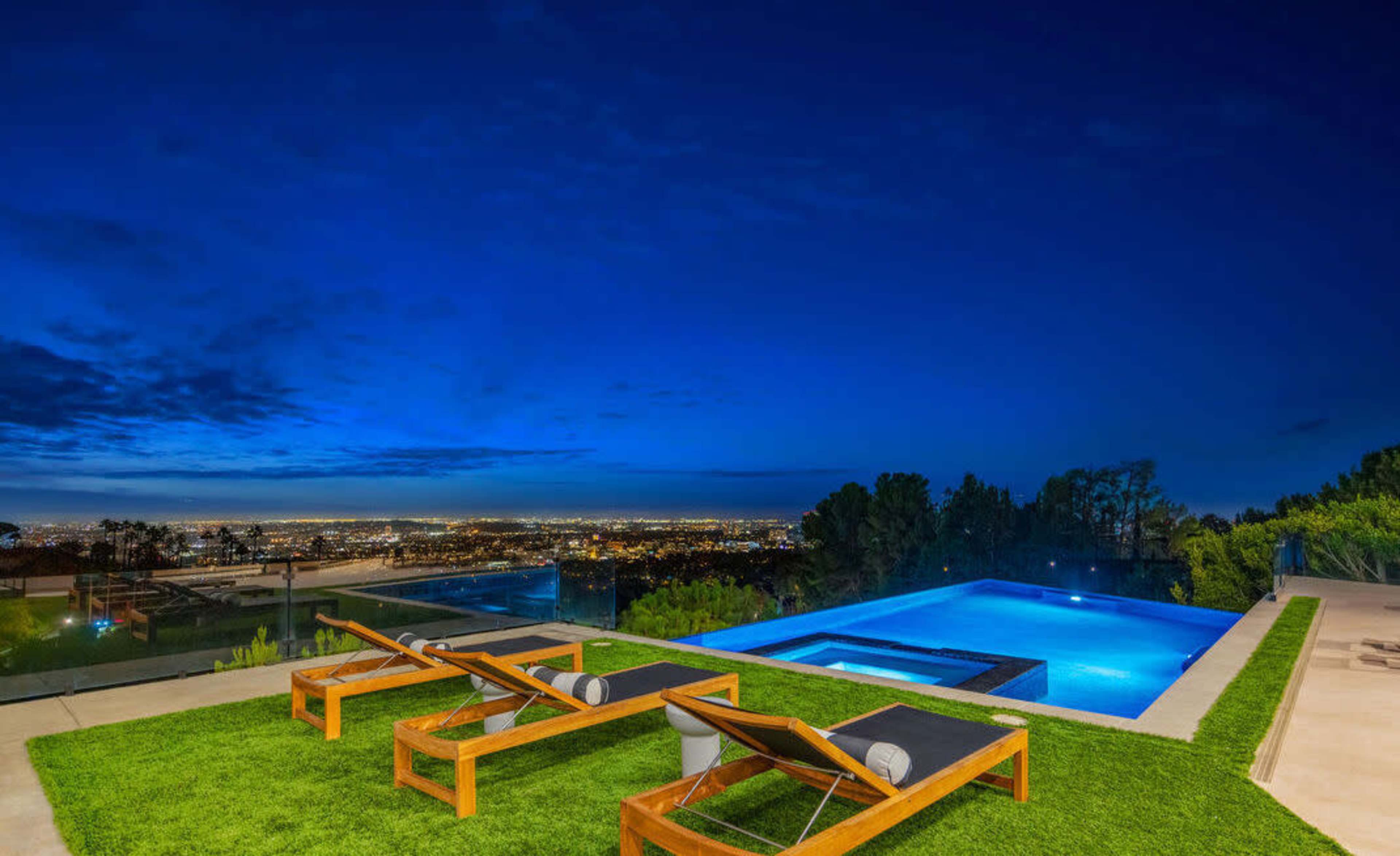 The scene features a modern pool area overlooking a city skyline at dusk, with lounge chairs positioned on green grass next to a glowing blue pool.