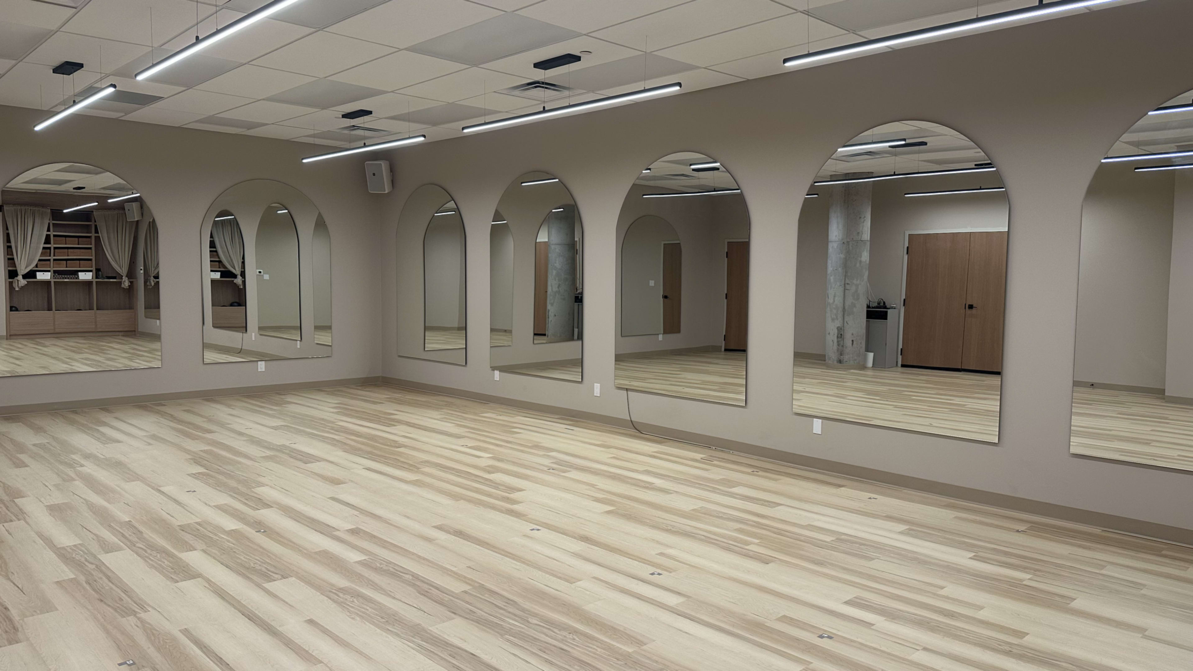 The image shows a spacious room with light wooden flooring and large arched mirrors on the walls, reflecting the neutral-colored interior.