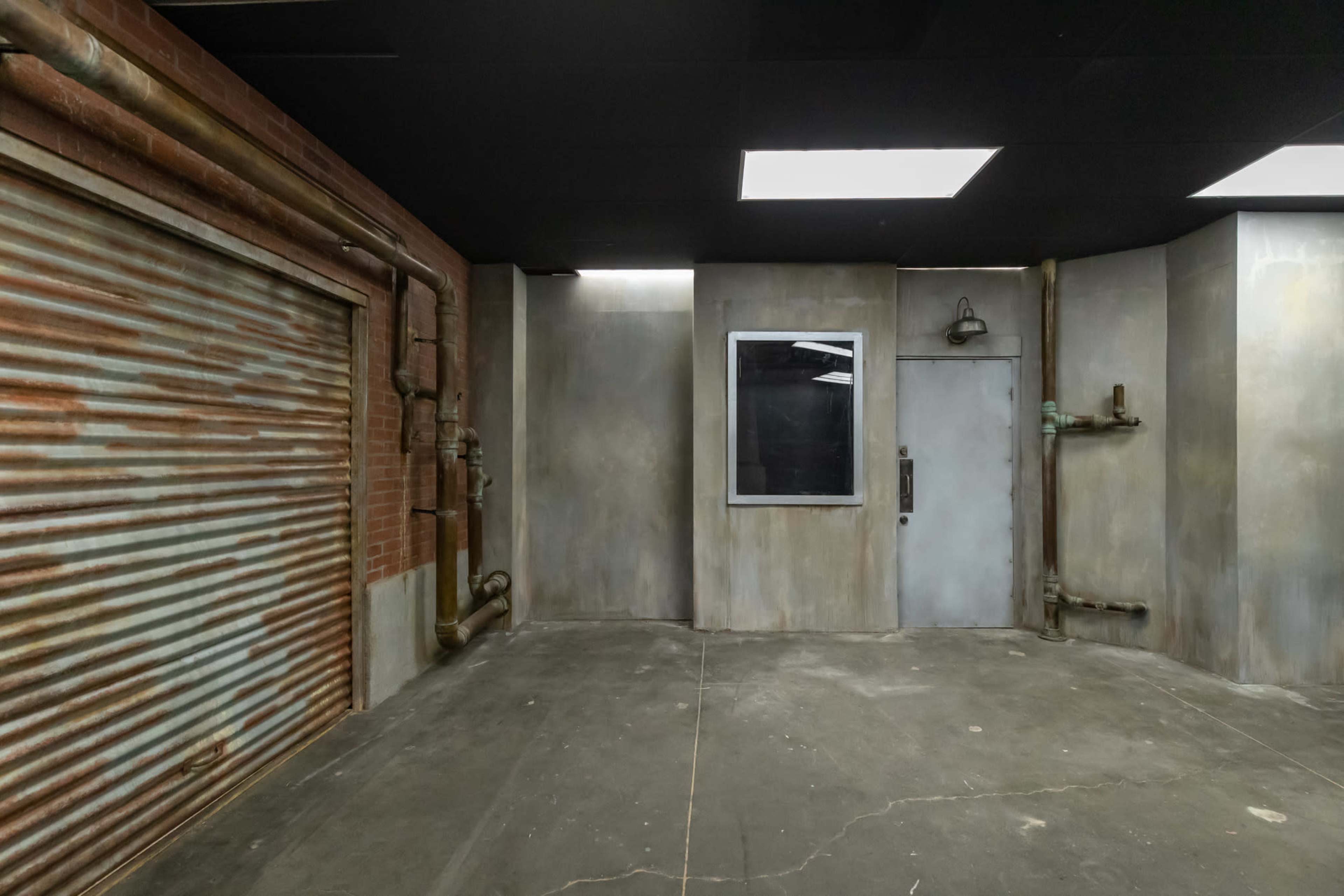 The image depicts an industrial-style room with a metal slat door, a small window, and exposed pipes on the walls.
