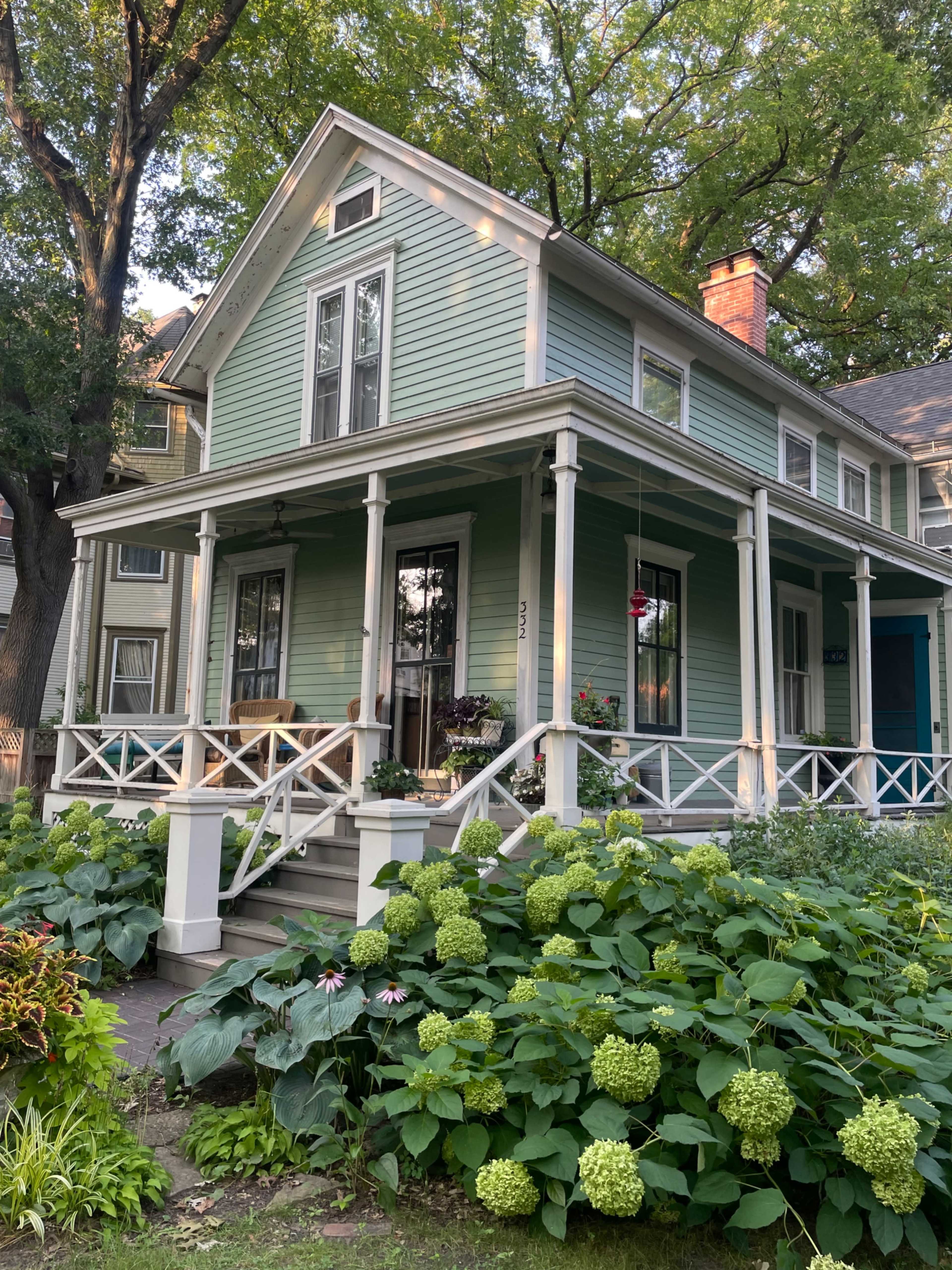 A two-story green house with a front porch is surrounded by lush greenery and flowering plants.