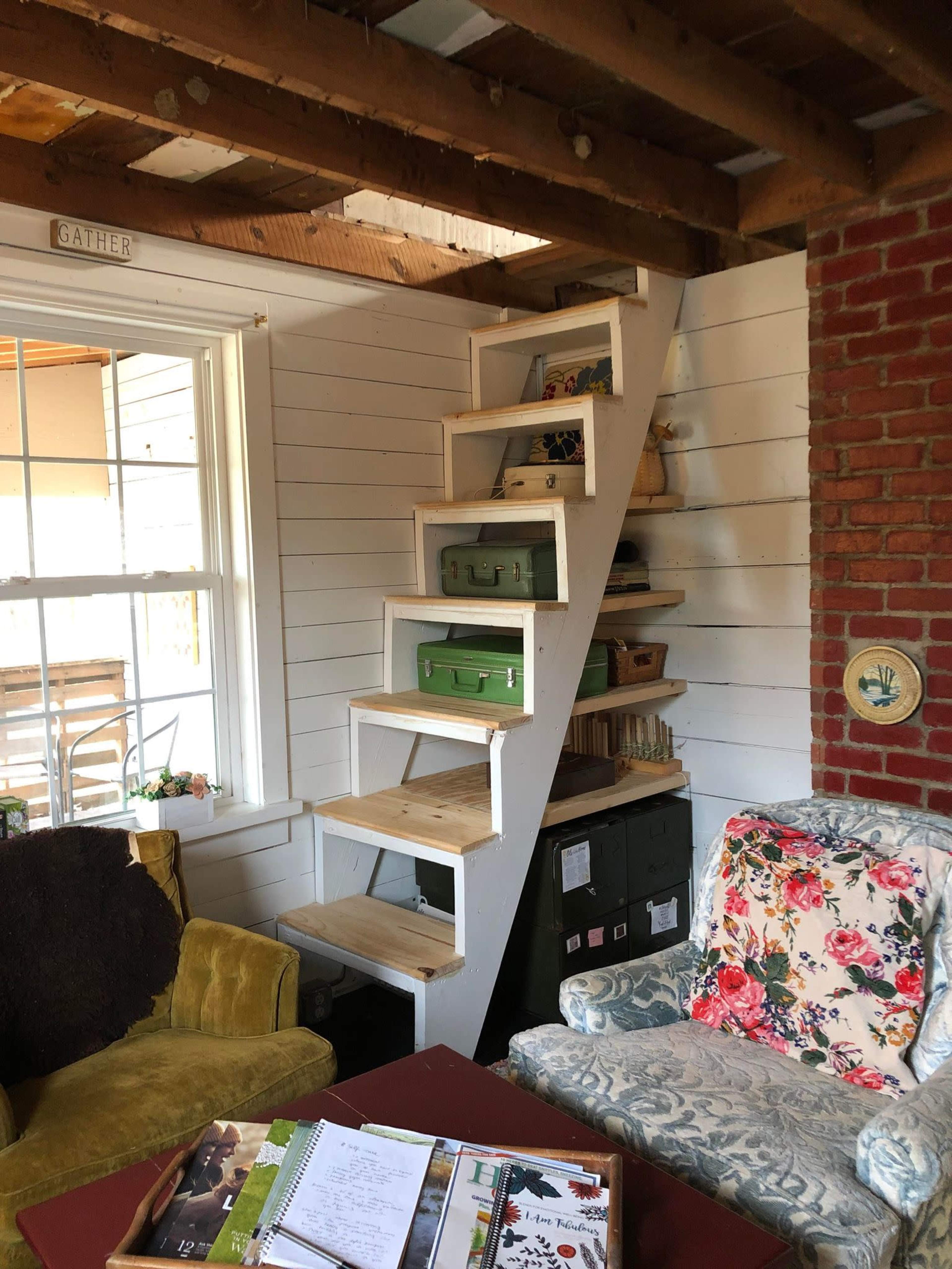 The image shows a cozy interior space with a white wooden staircase leading to an upper level, surrounded by colorful furniture and shelves.