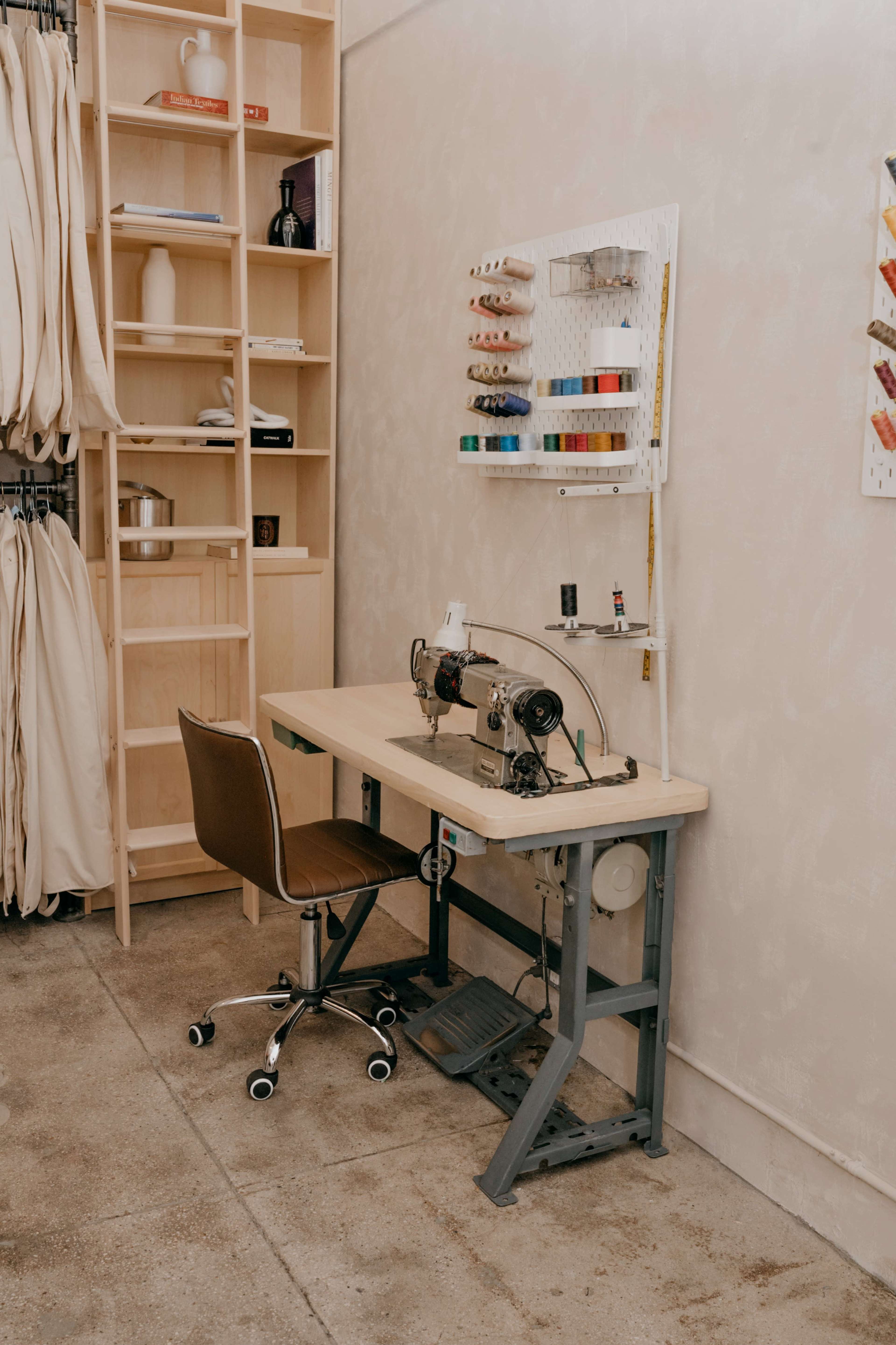 The image shows a sewing workstation with two sewing machines on a wooden table, accompanied by a chair and a wall-mounted organizer for threads.