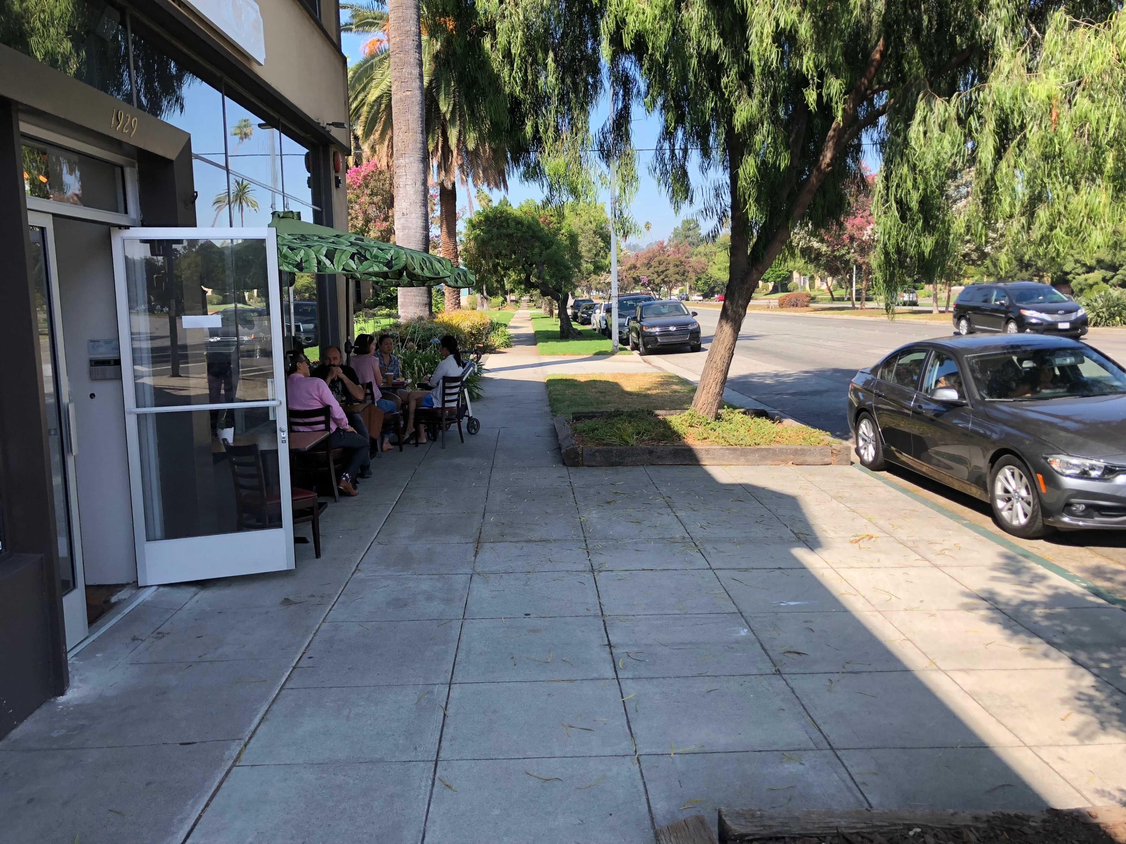 The image shows a sidewalk next to a street with several parked cars, a dining area with patrons outside a restaurant, and palm trees in the background.