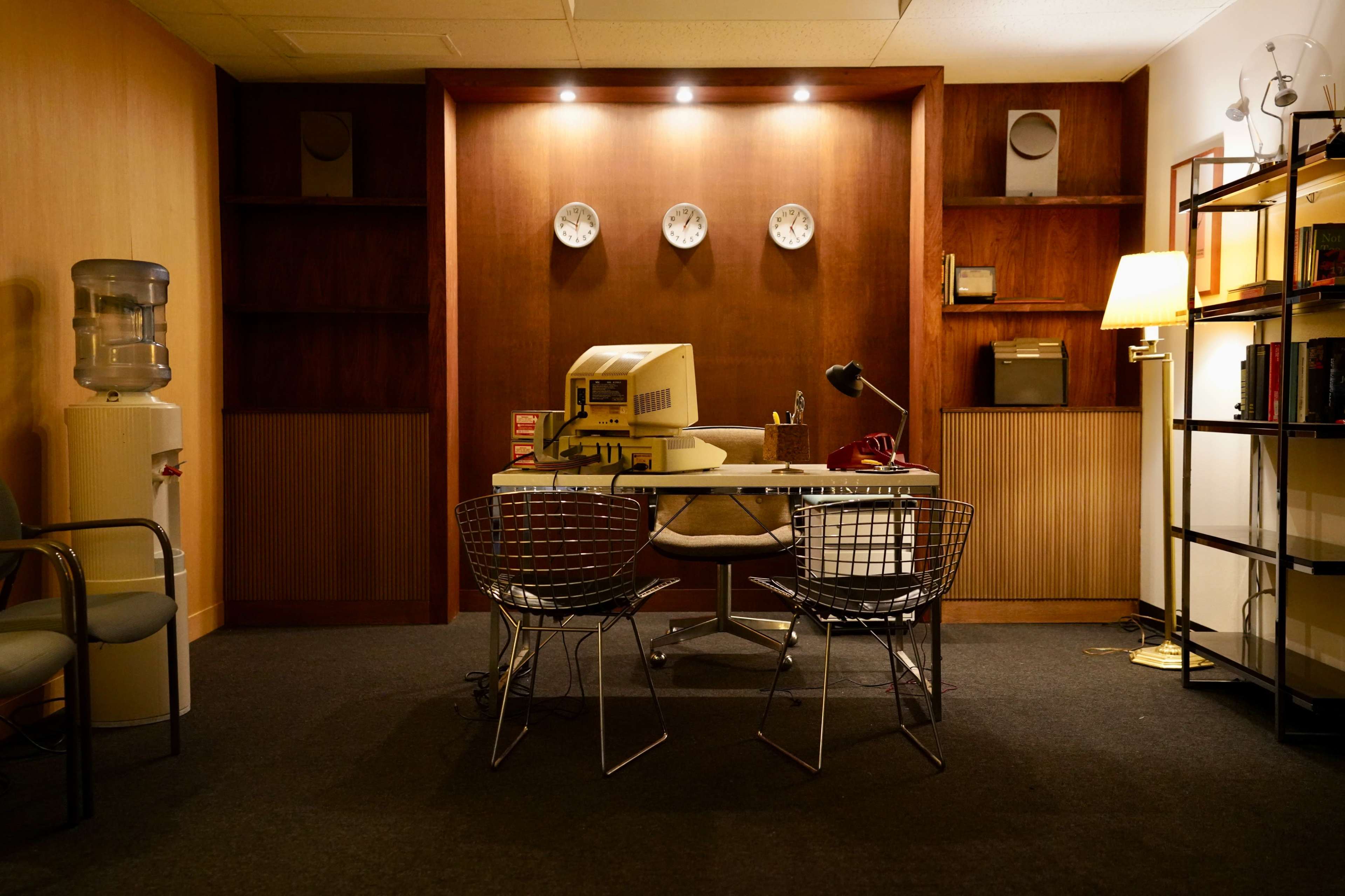 The image shows a vintage office space featuring a desk with an old computer, two chairs, a water cooler, and various clocks on the wall.