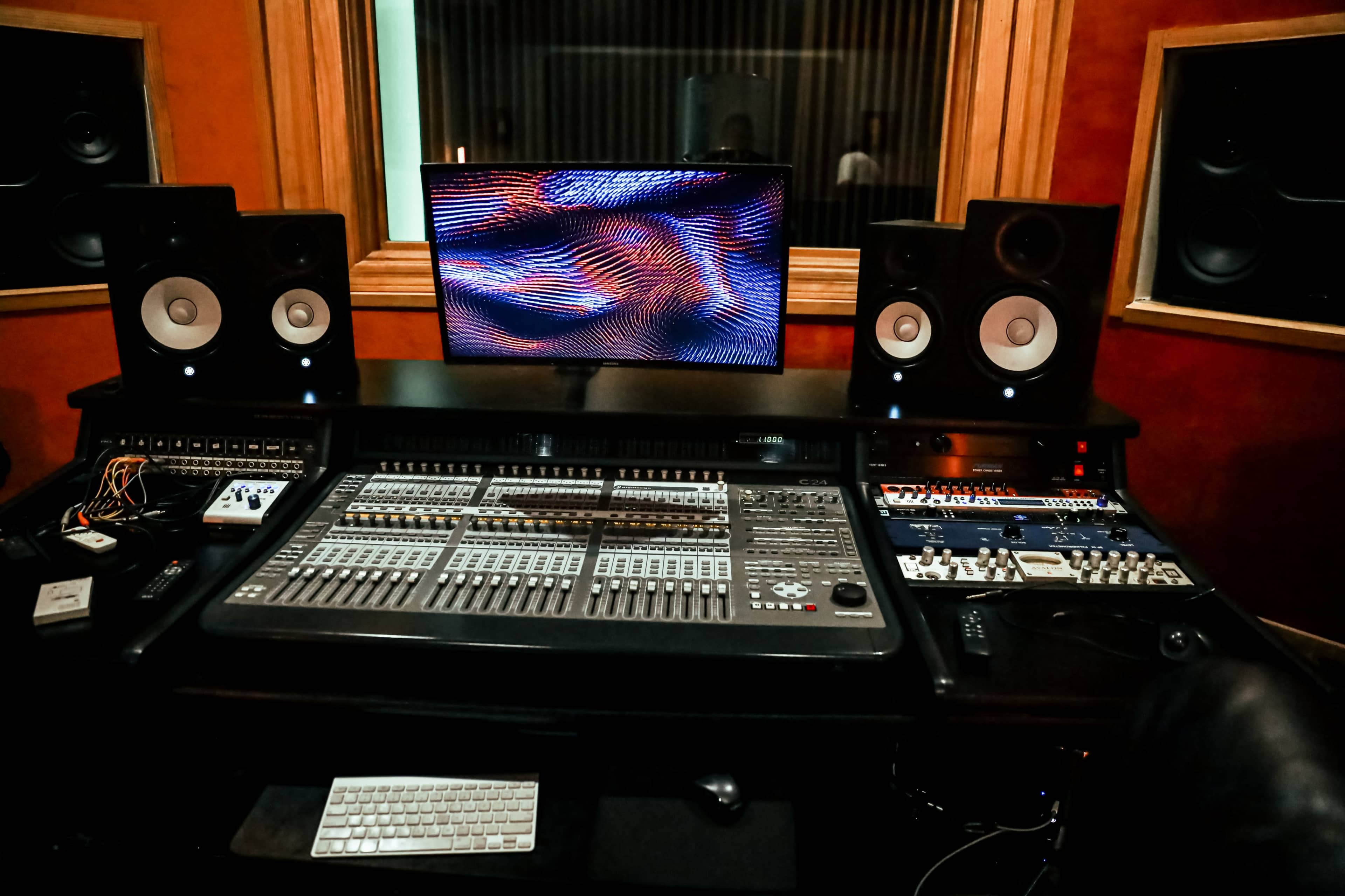 The image shows a recording studio setup featuring a mixing console, two monitors, and various audio equipment in a soundproofed environment.