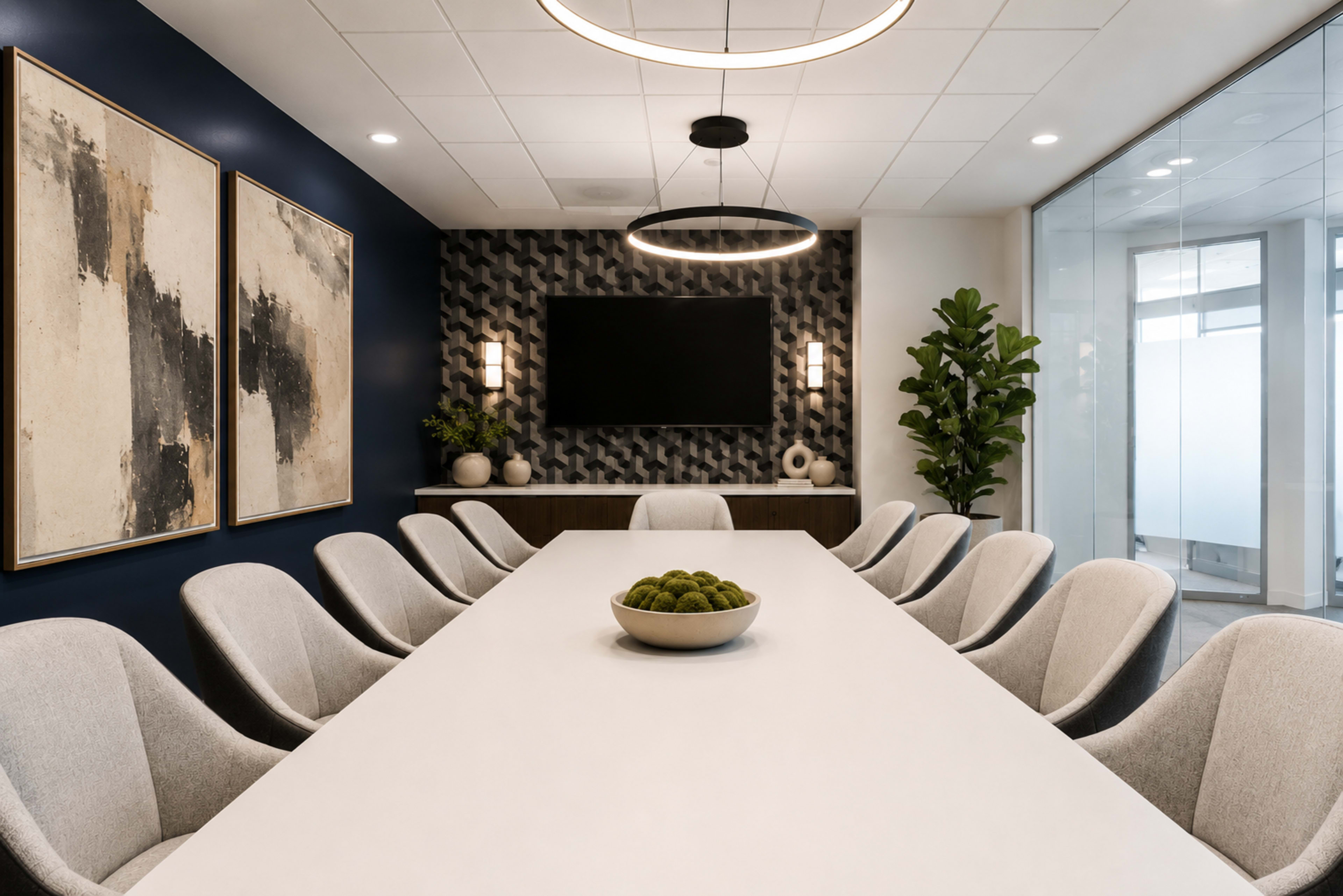 A spacious conference room features a long white table surrounded by gray chairs, with artwork on the dark blue walls and a TV mounted on the accent wall.